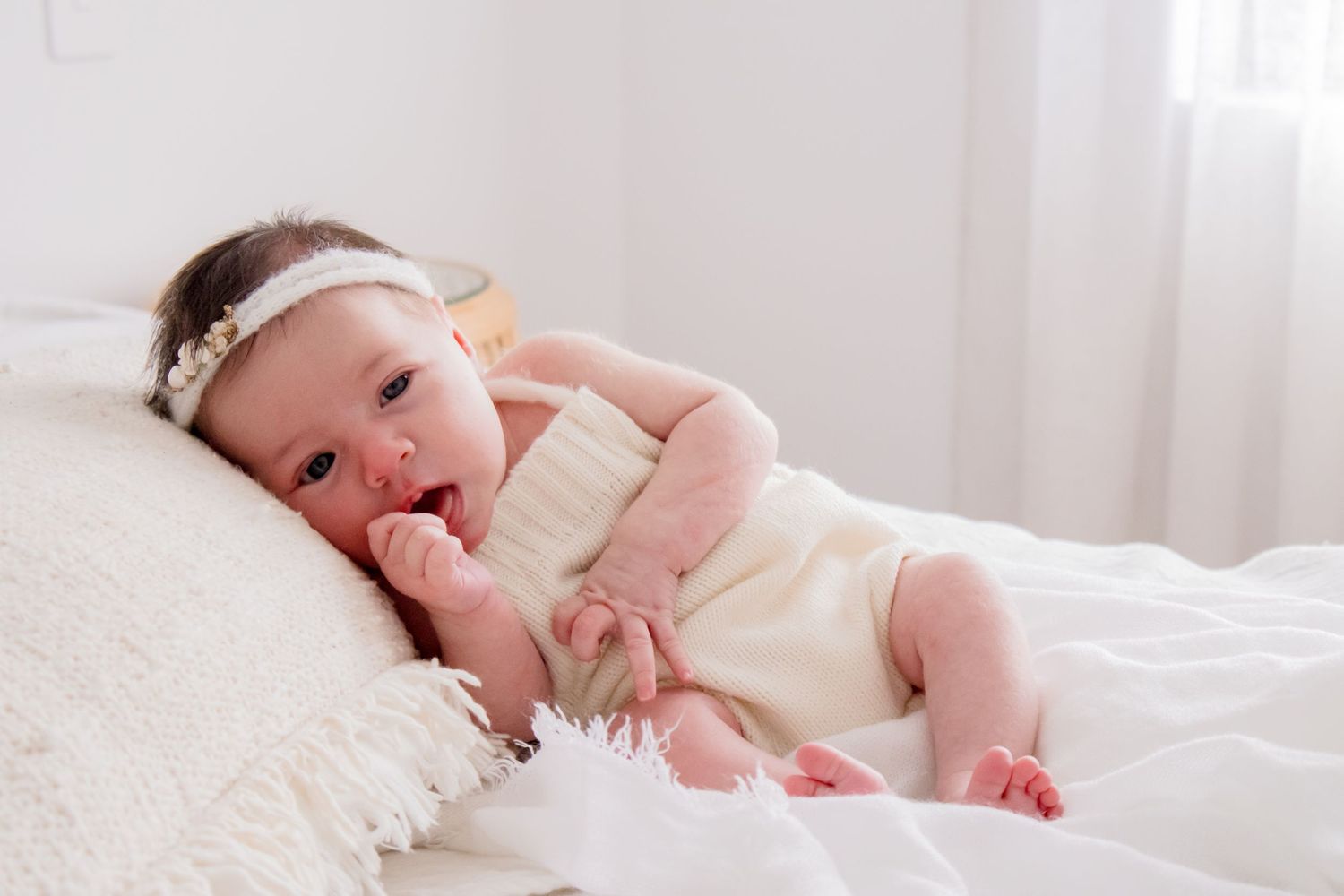 Baby in cream romper lying on white pillow with hand near mouth.