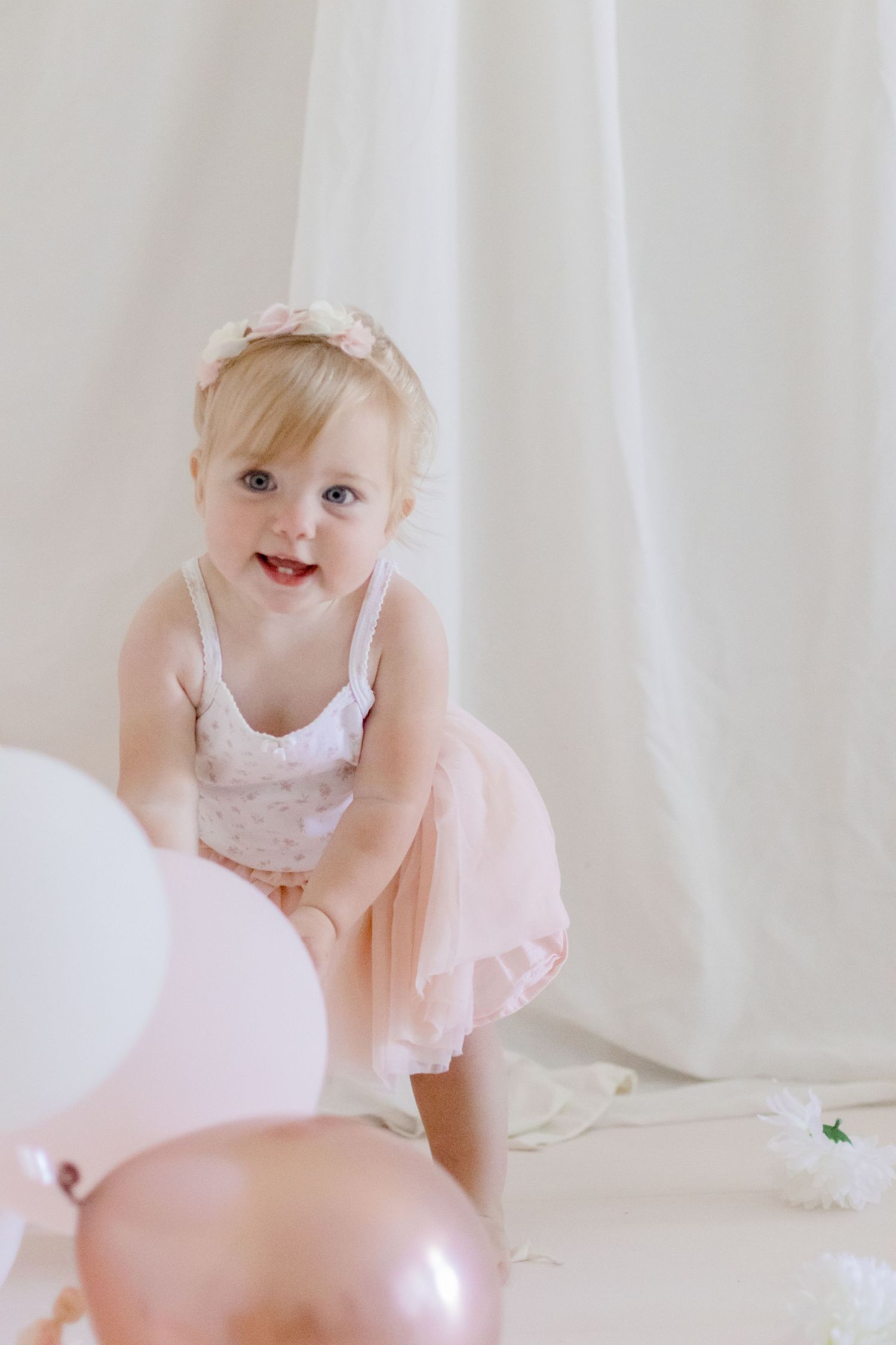 Toddler in a flowy pink dress playing with balloons against a white background.