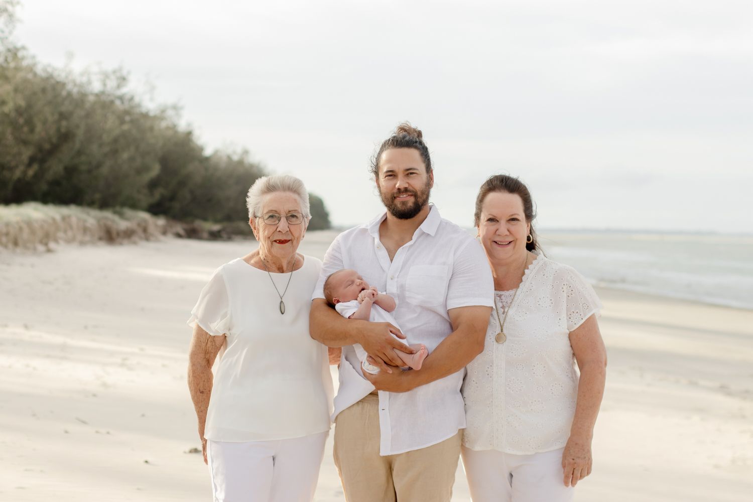 Three generations pose together on a beach in matching white outfits.
