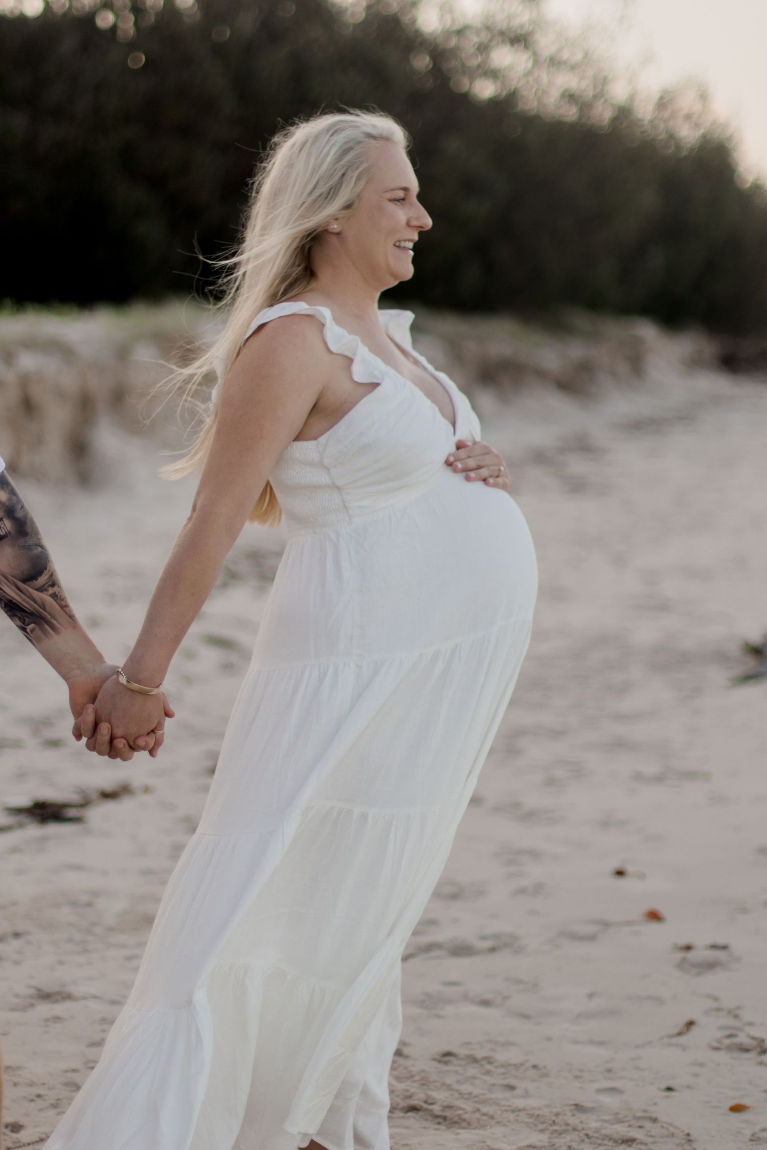 A pregnant woman in a white maxi dress smiles during her beach maternity session.