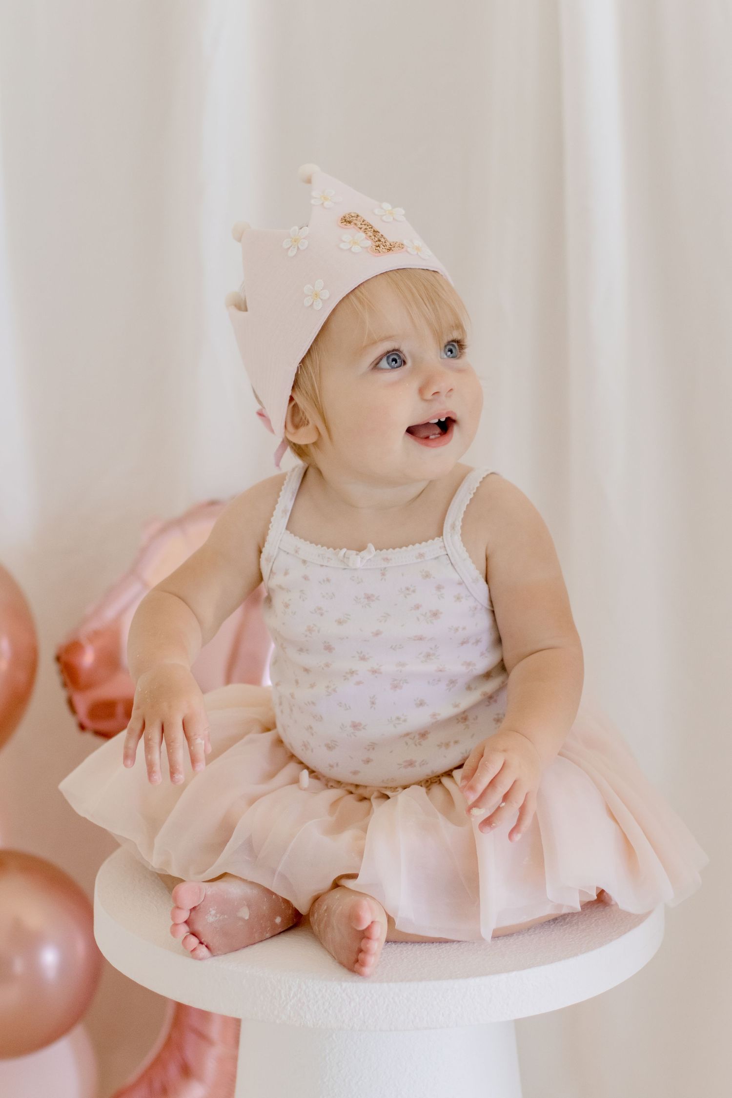 Baby in a light pink outfit and crown laughs while sitting on a white pedestal near rose gold balloons.