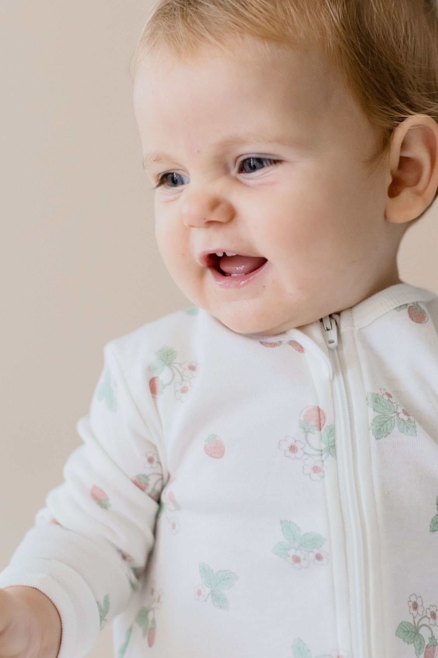 Baby wearing white floral zip-up outfit smiles against beige background.
