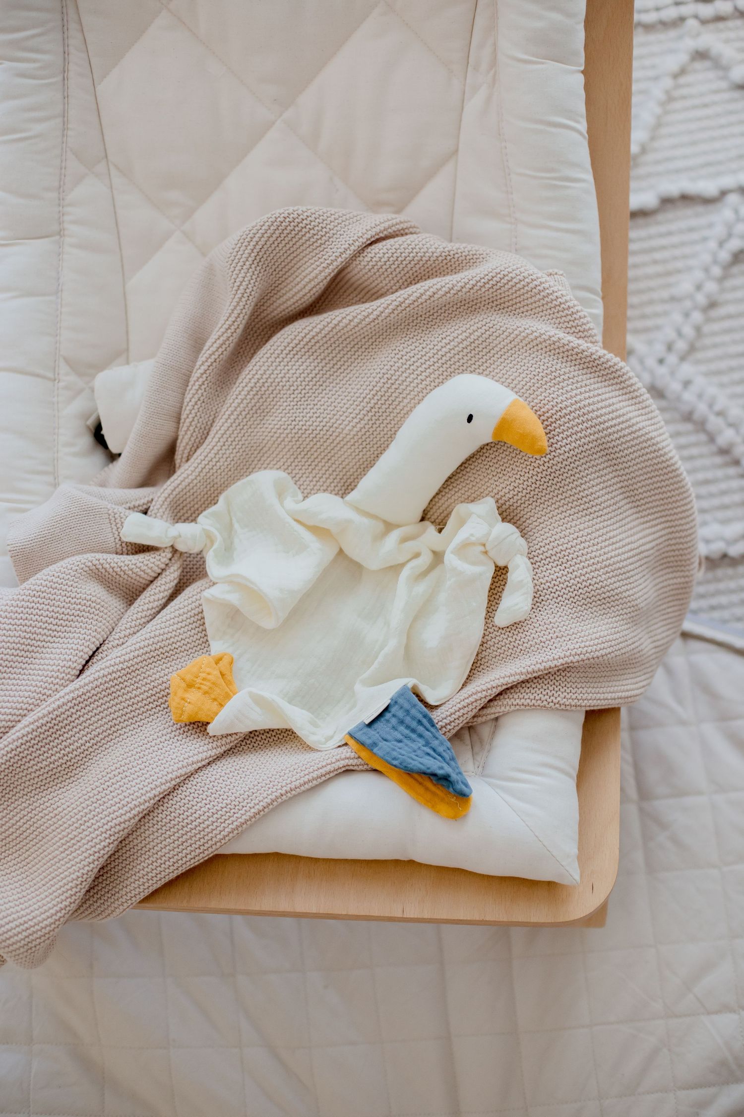 White duck plush toy with blue boots resting on beige knit blanket.