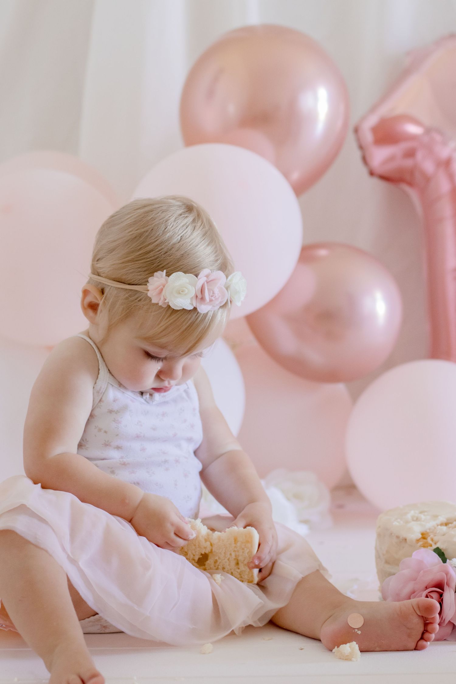Small child in tulle dress with rose headband examining food against pink balloon background.