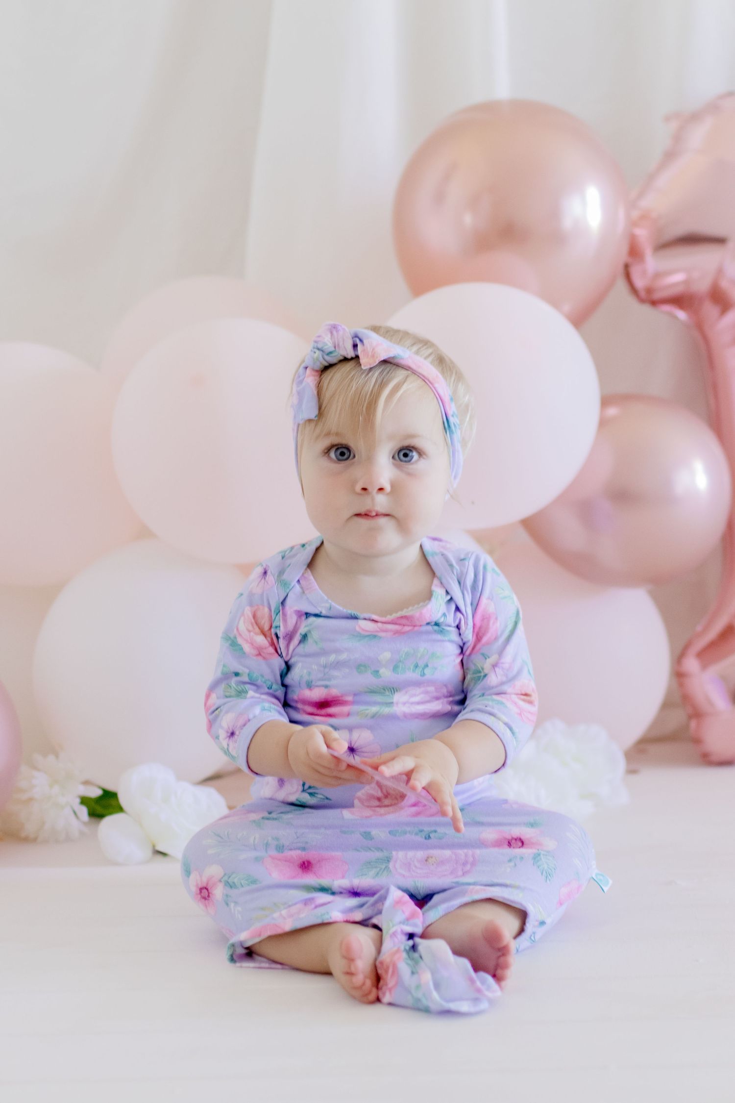 A watercolor floral romper paired with a bow headband against pink balloon decorations.