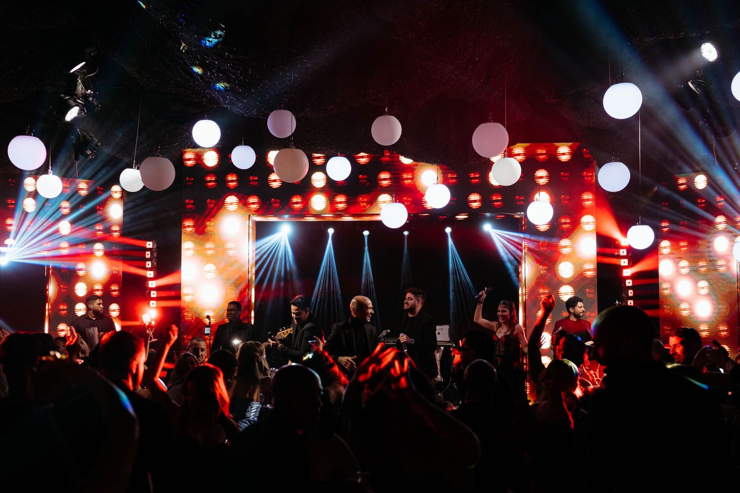 Live concert stage with glowing white balloons, vibrant red lights, and a crowd enjoying an energetic night performance.