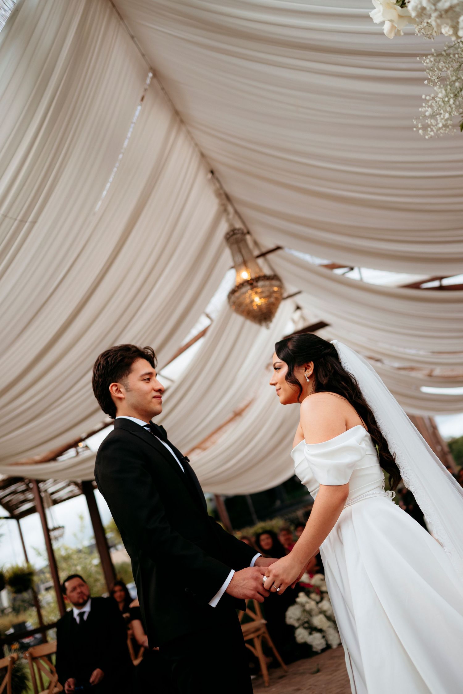 Bride and groom hold hands at wedding altar under draped white fabric canopy.