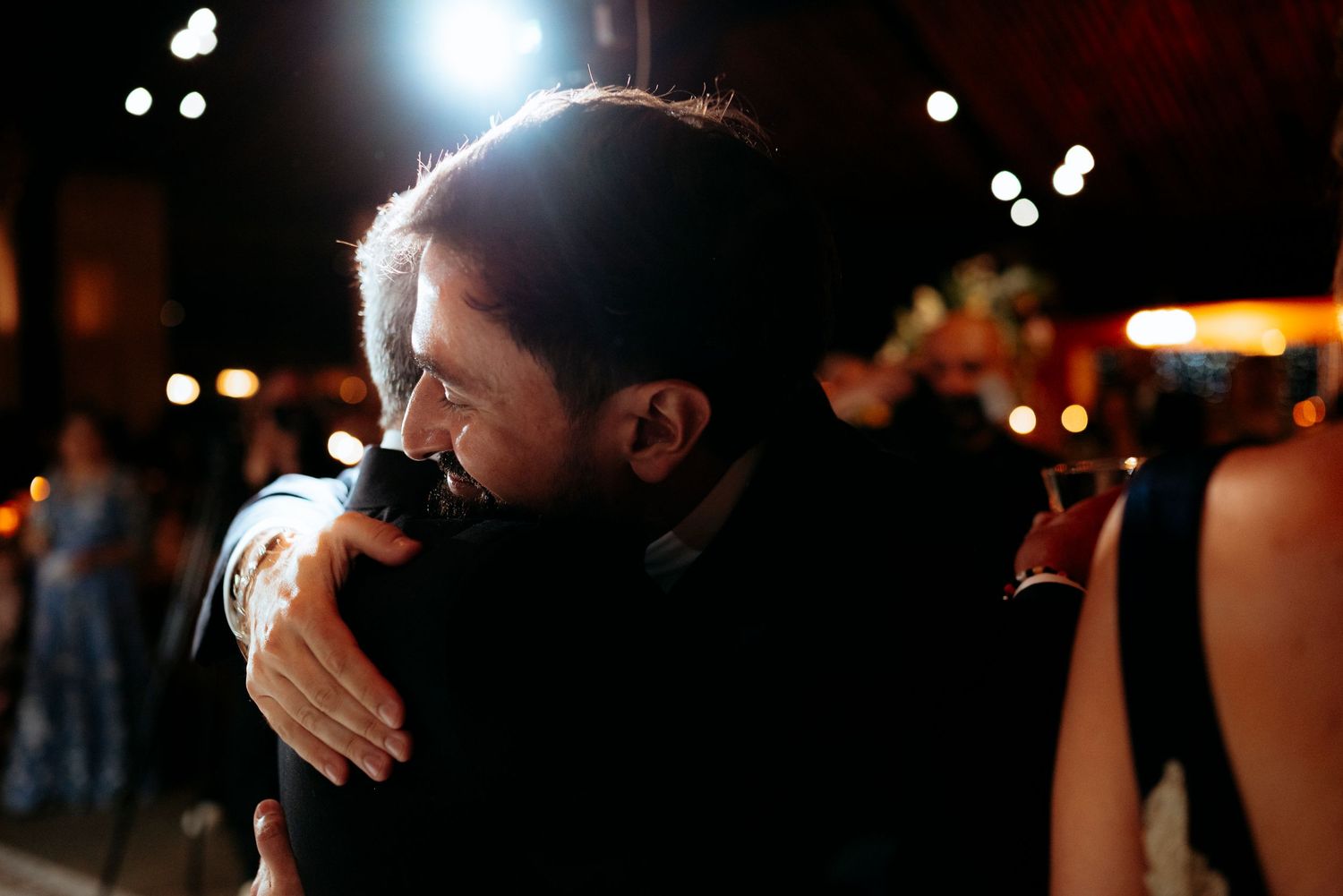 Two people sharing a warm embrace at a nighttime outdoor event with bokeh lights in the background.