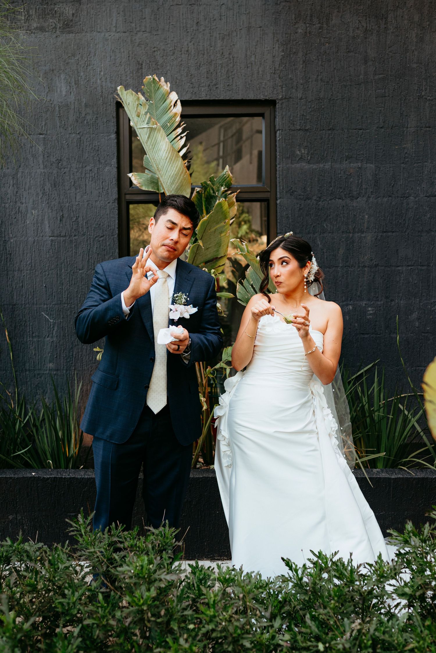 Bride and groom blowing bubbles in wedding attire outside against a dark wall with tropical plants.