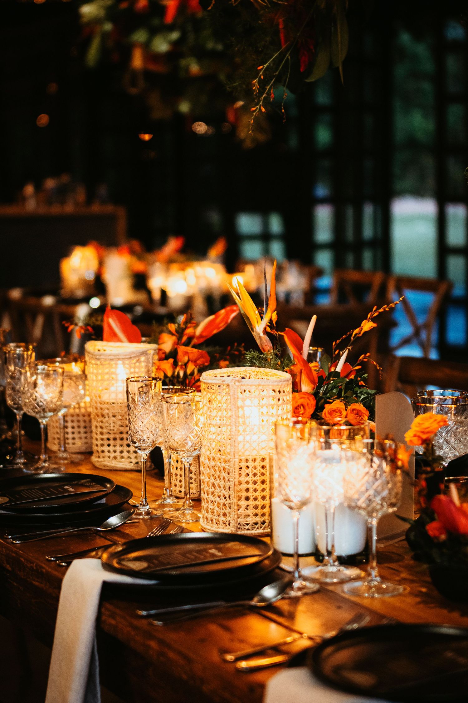 Elegant wedding reception table with glowing candles, crystal glasses, black plates, and vibrant orange floral centerpieces on rustic wood.
