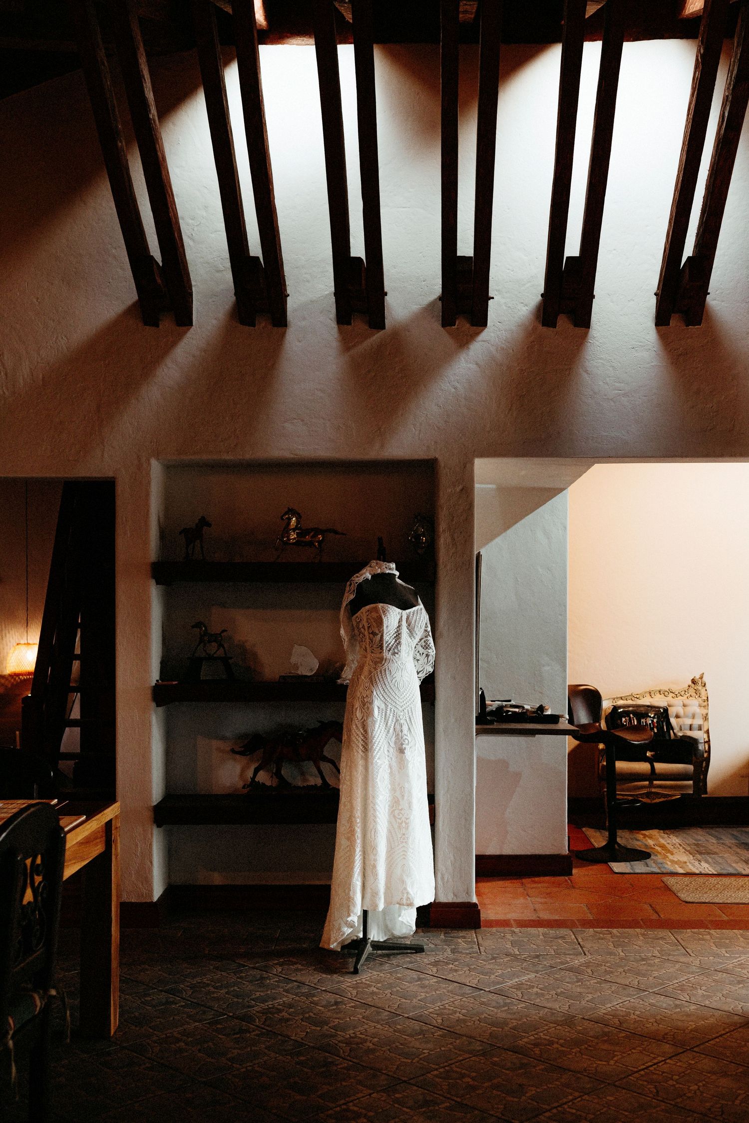 A white wedding dress hangs in a dimly lit interior room with exposed wooden beams and brick flooring.