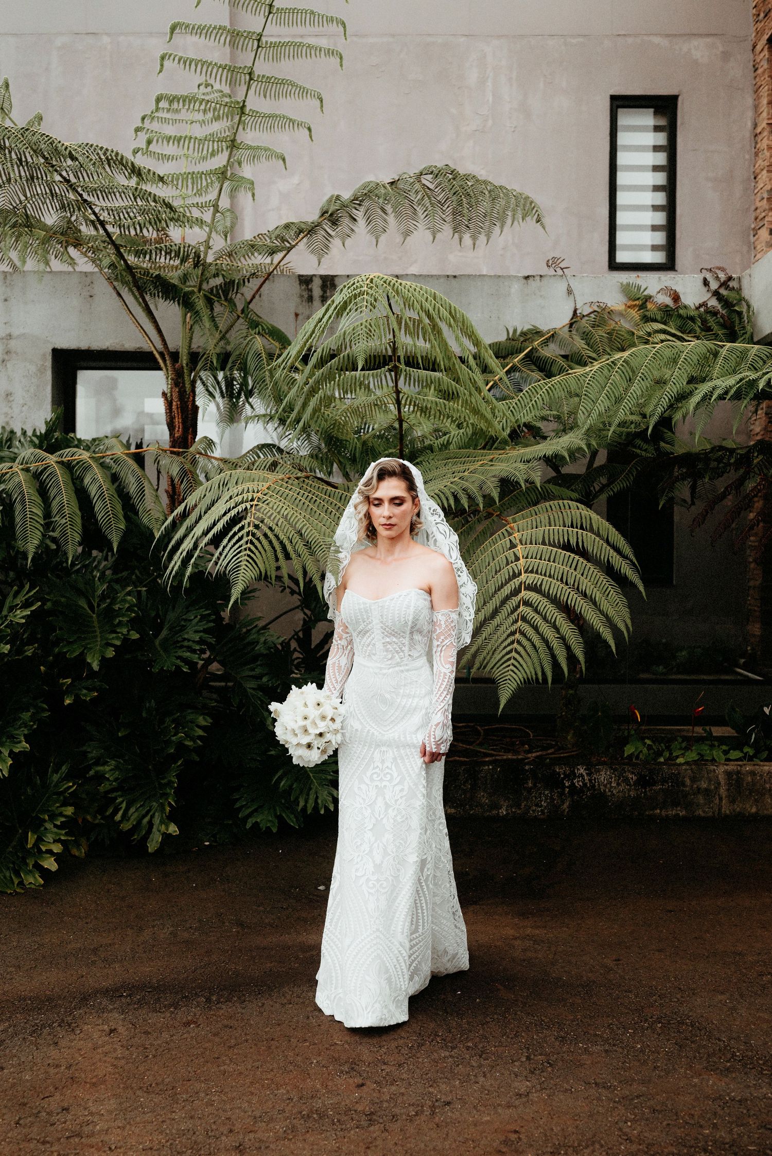 A bride in lace stands elegantly among large tropical ferns against a white wall.