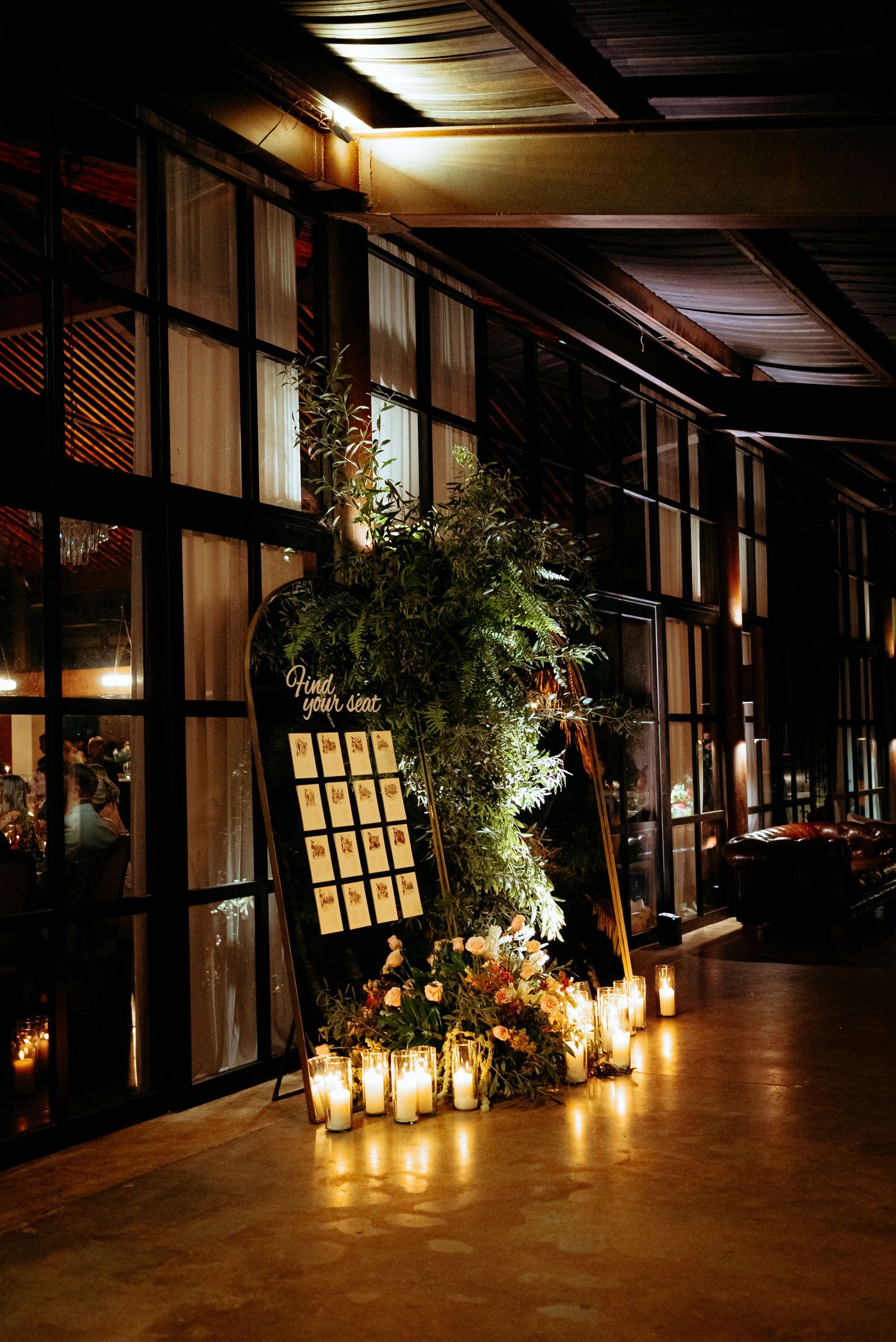 Elegant wedding seating chart display with lush greenery, floral arrangements, and glowing candles in a dark venue.