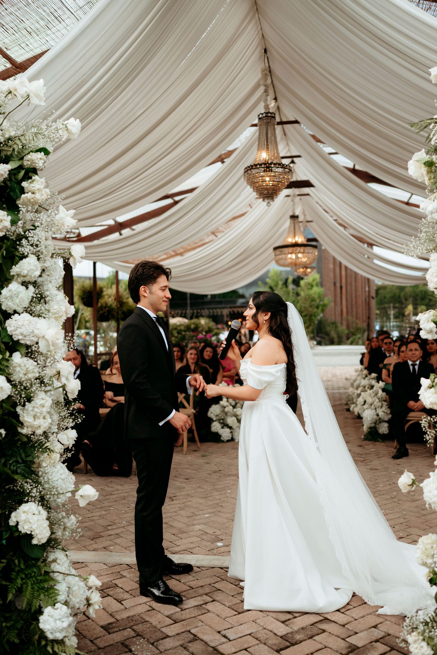 Bride and groom exchange vows under elegant white draped canopy with crystal chandeliers and white floral arrangements.