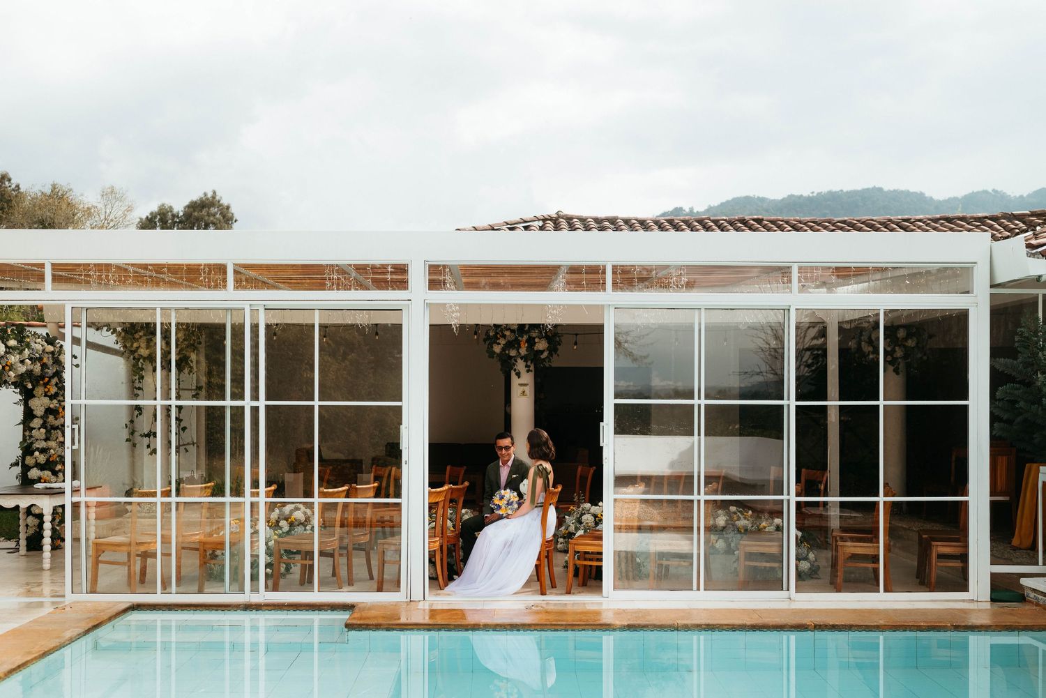 Bride and groom share a romantic kiss by a pool at an elegant outdoor wedding venue with glass walls.