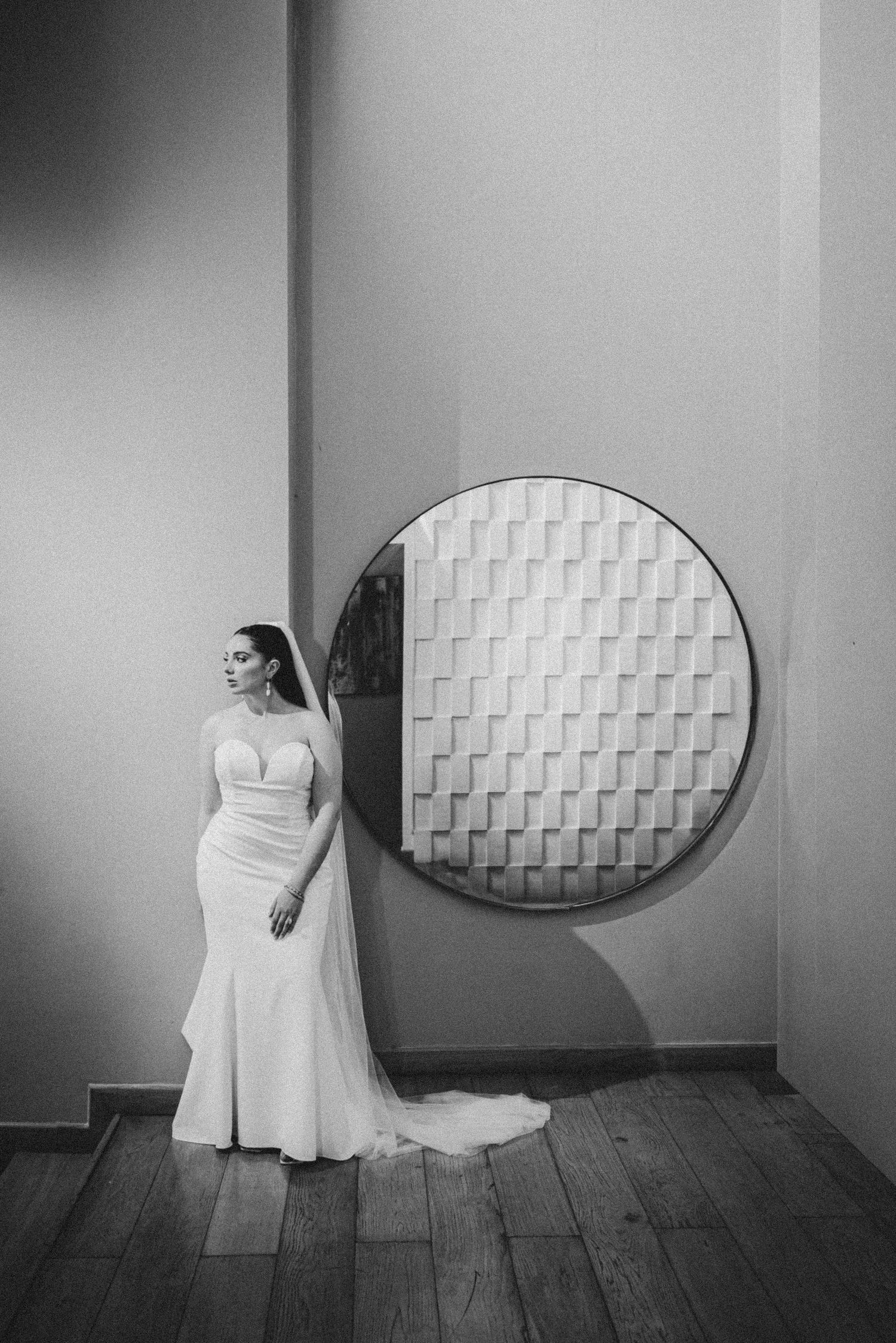 Bride in a fitted white gown with veil stands beside a circular decorative mirror in a black and white photo.