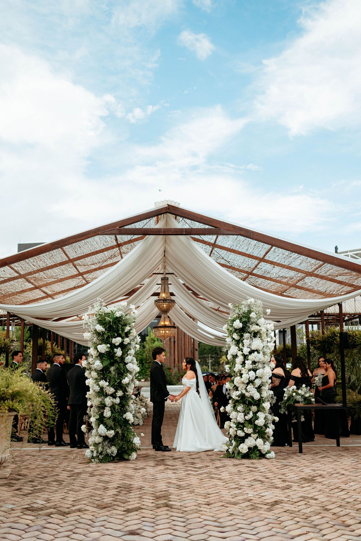 Bride and groom exchange vows under a white draped pergola adorned with tall white floral columns at an outdoor wedding ceremony.