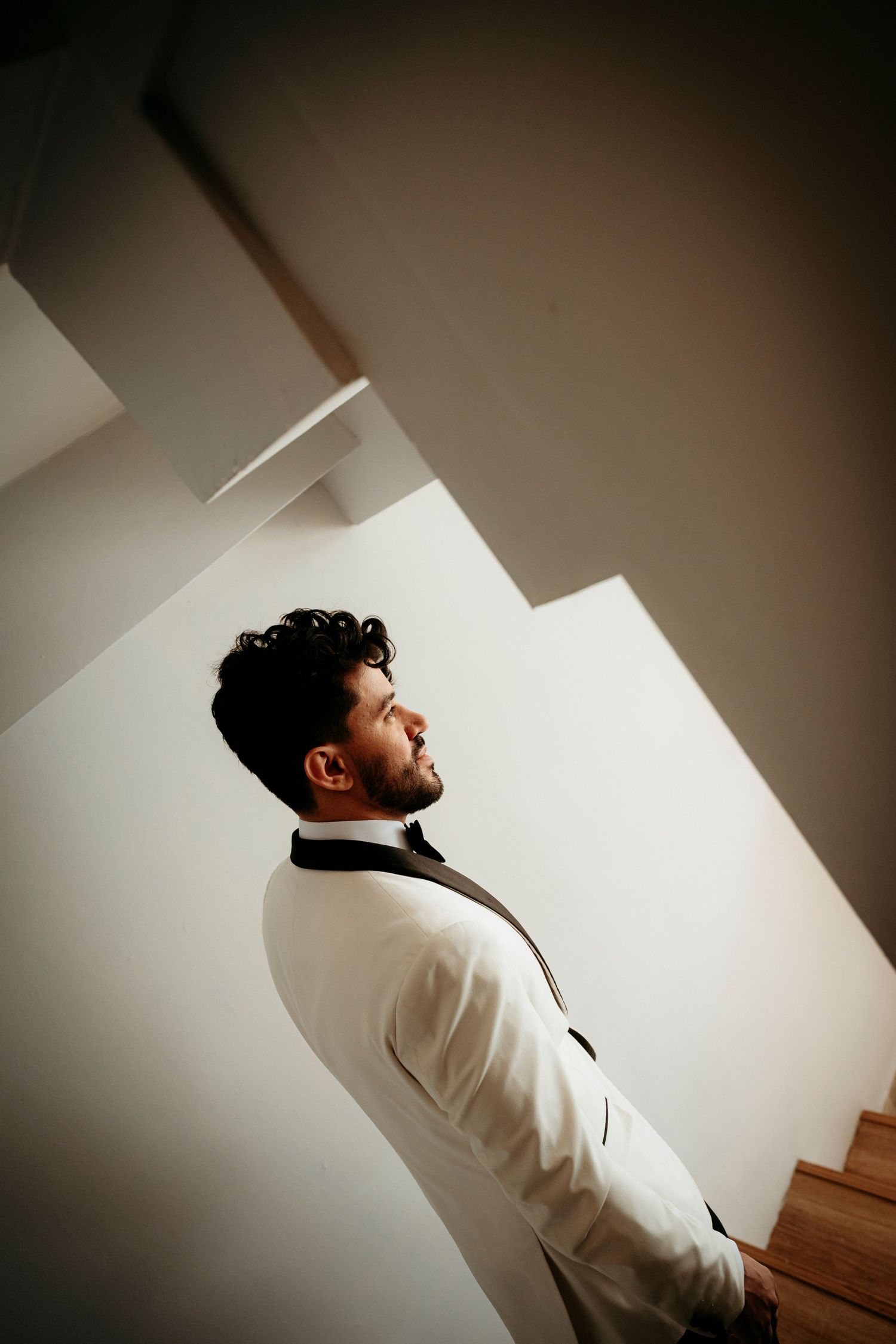A bearded man in a white blazer gazes upward while standing near a staircase in a modern white interior.