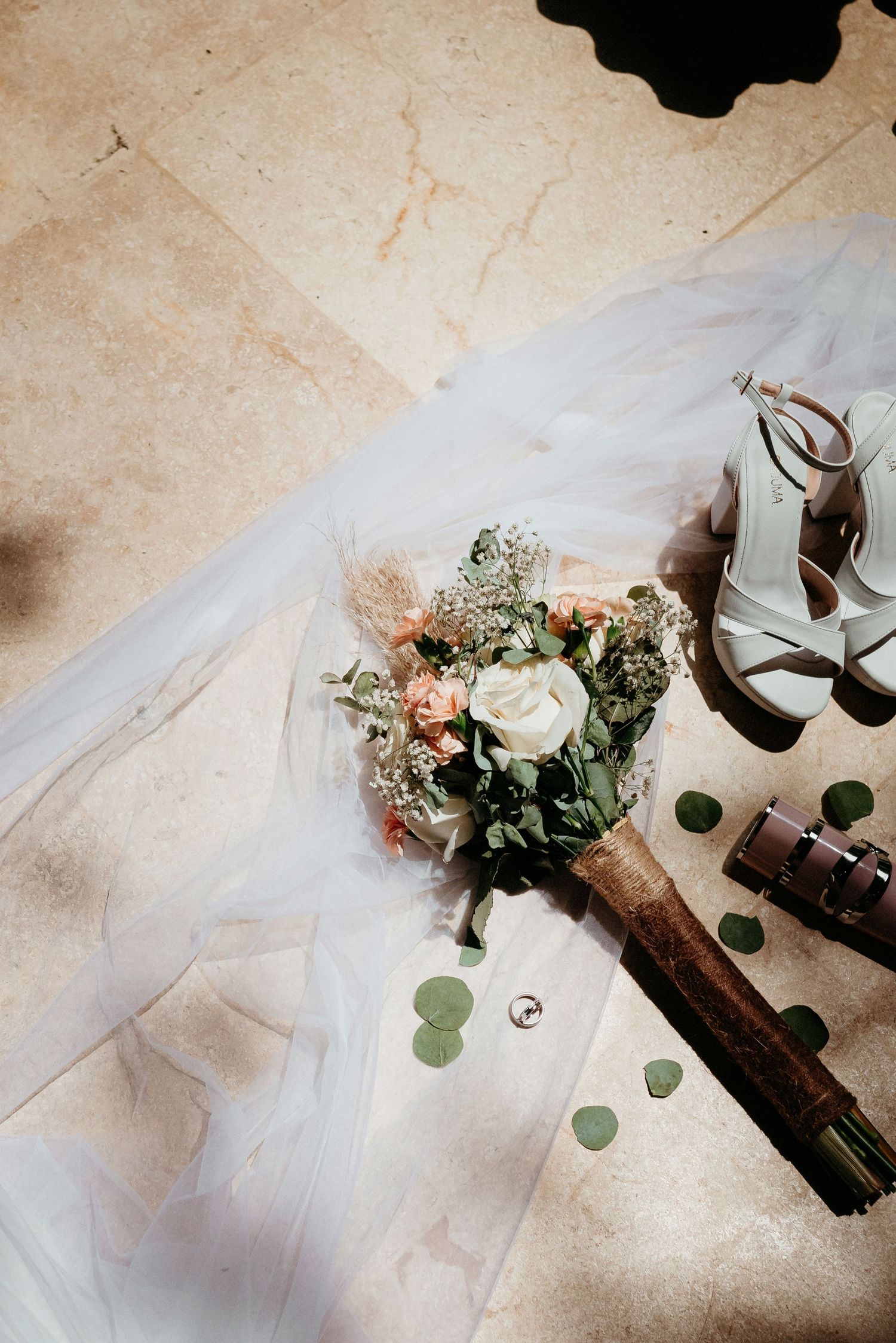A bridal bouquet of white roses and greenery lies next to wedding shoes and veil on a light stone floor.