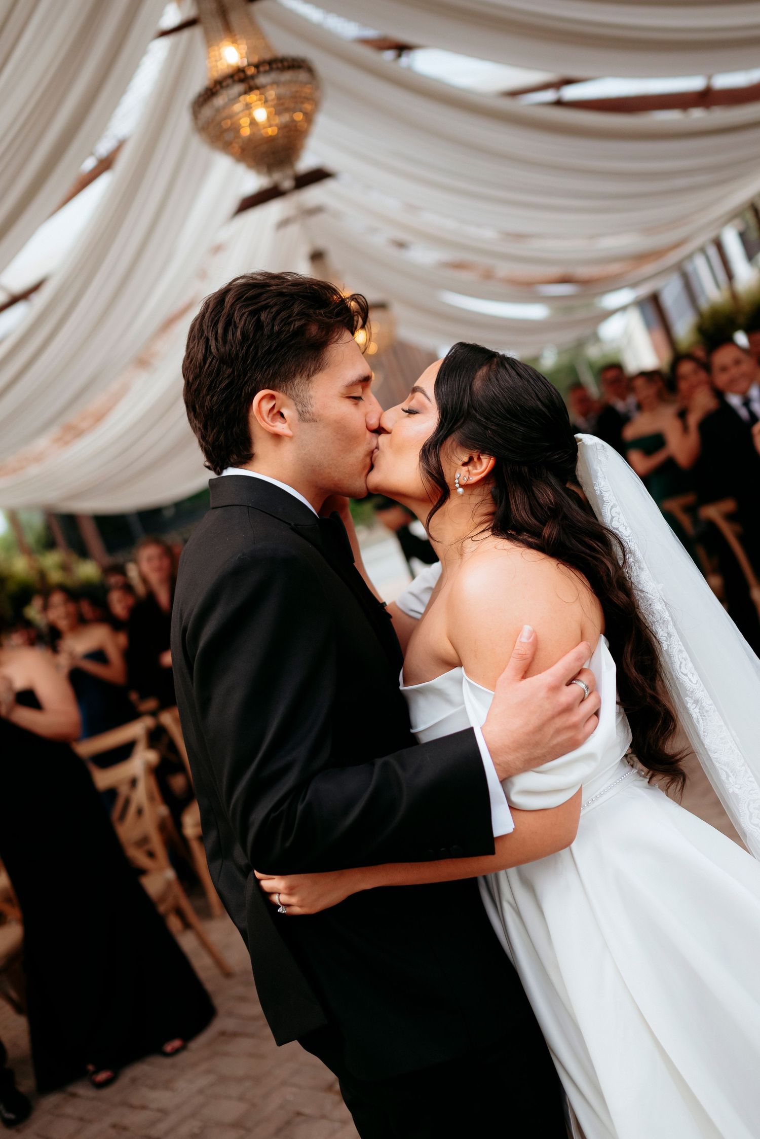 Bride and groom share their first kiss under elegant draped fabric ceiling with chandelier at wedding ceremony.