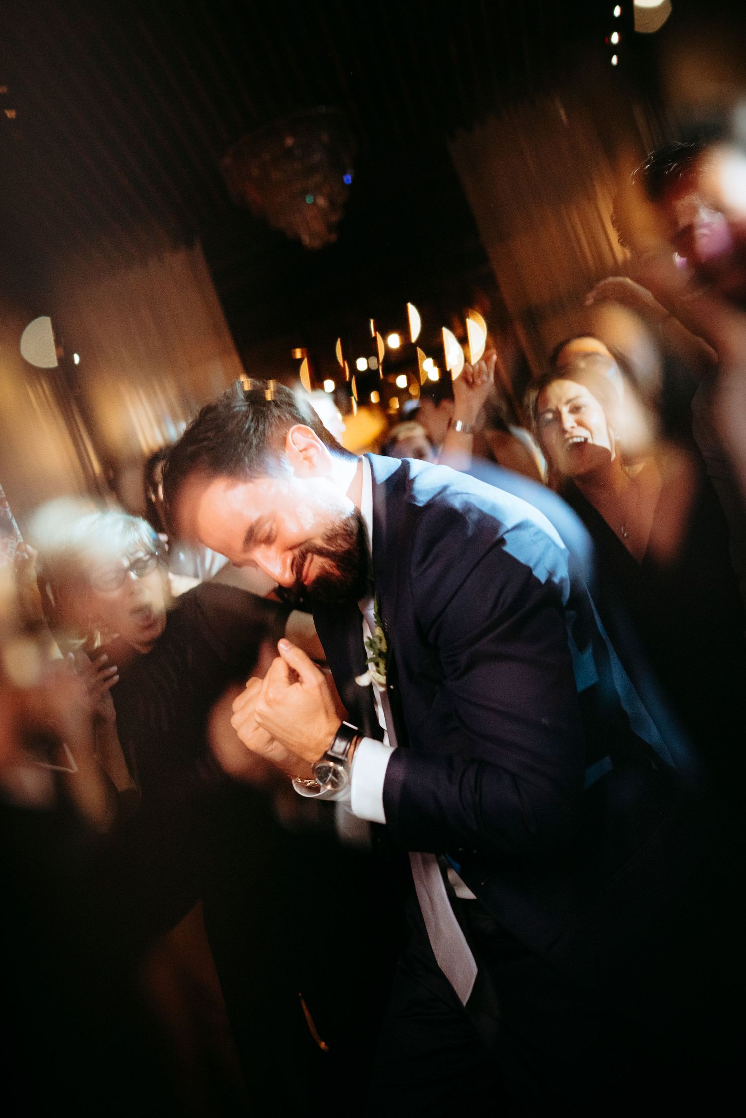 Groom dipping partner on dance floor surrounded by cheering wedding guests in dimly lit reception venue.