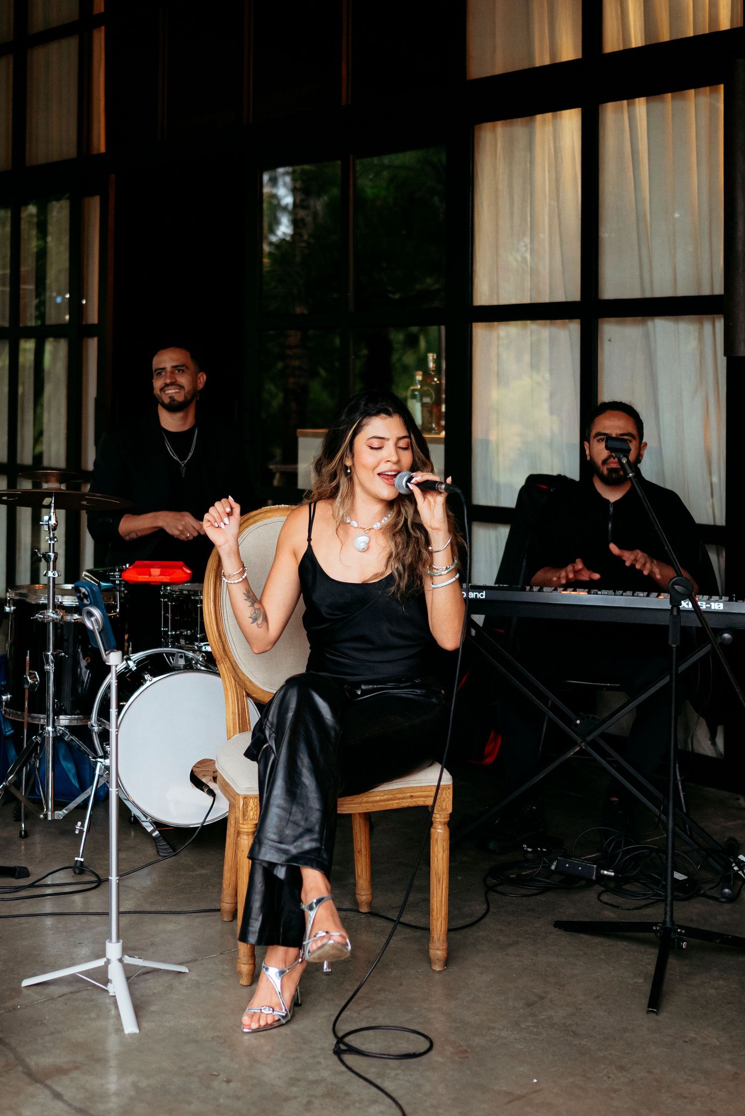 Female singer performing live with band, wearing black outfit, seated with microphone at elegant indoor venue.