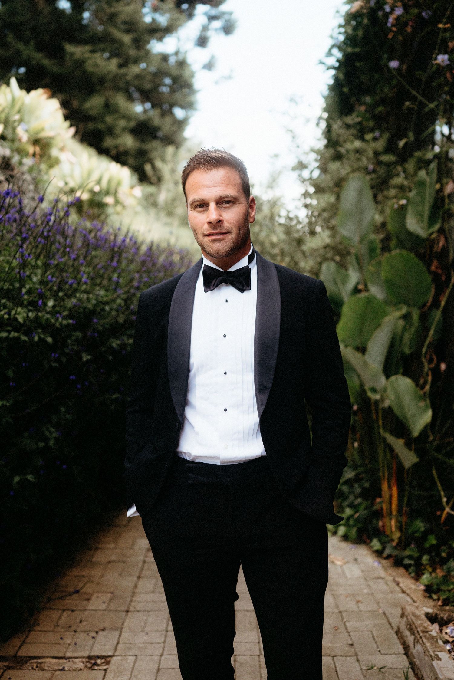 A groom in a classic black tuxedo and bow tie stands on a garden path surrounded by flowering plants and greenery.