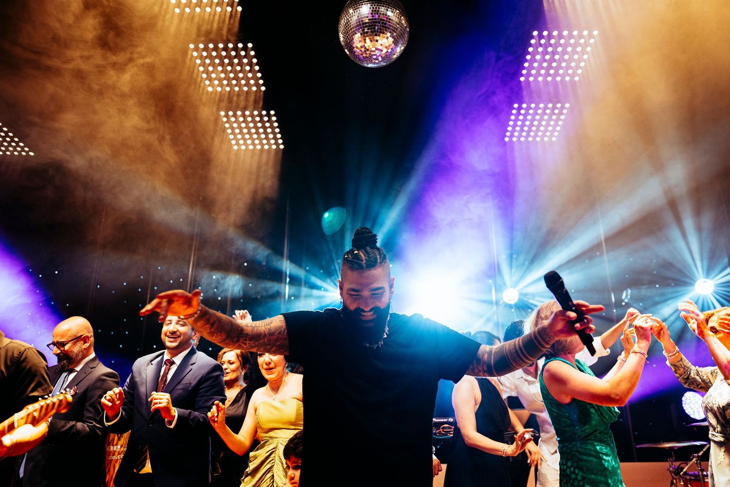 Energetic dance party scene with performer under disco ball, colorful stage lights, and excited crowd in formal attire.