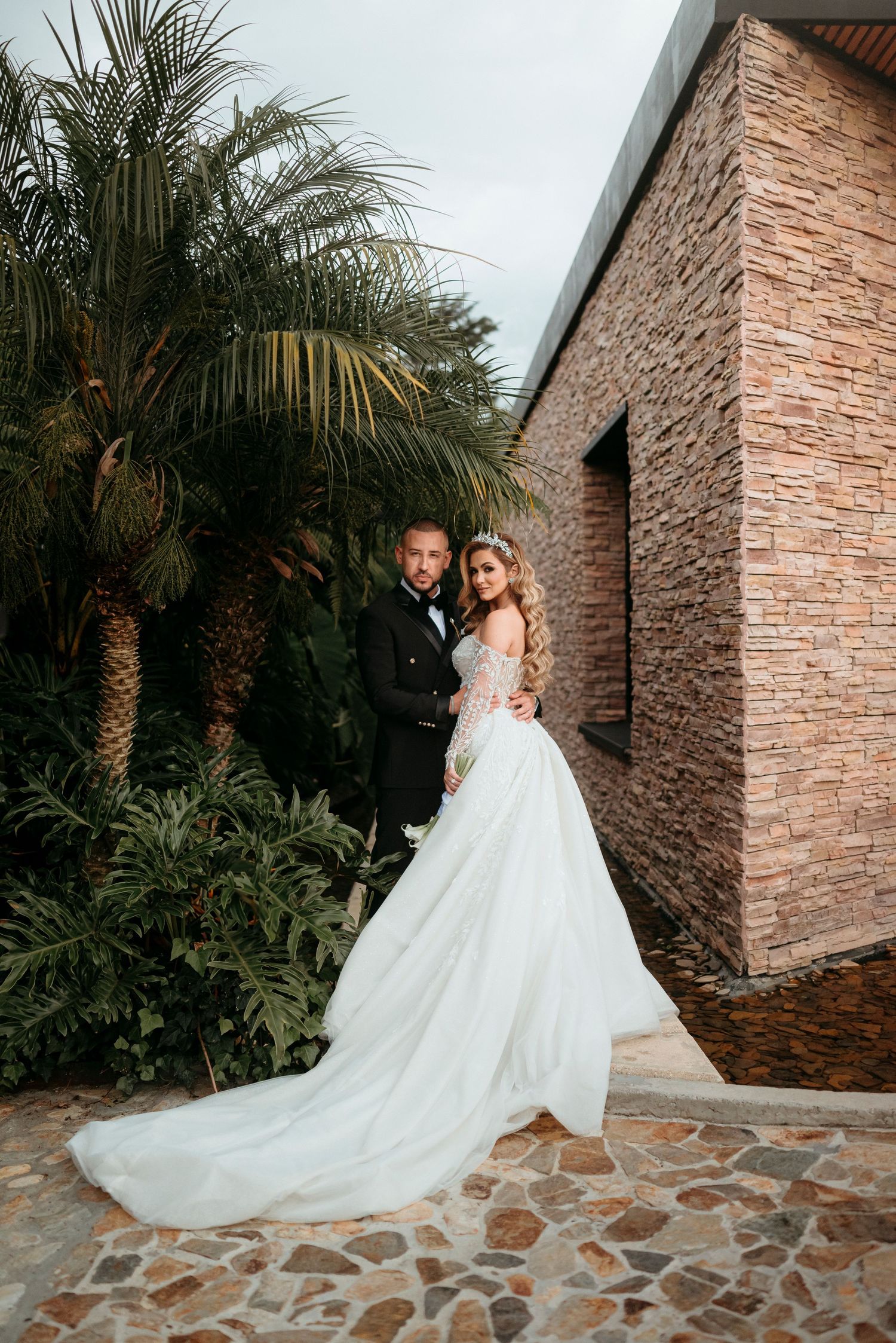 Bride in elegant white gown with long train stands with groom in tuxedo near tropical palms and stone building.