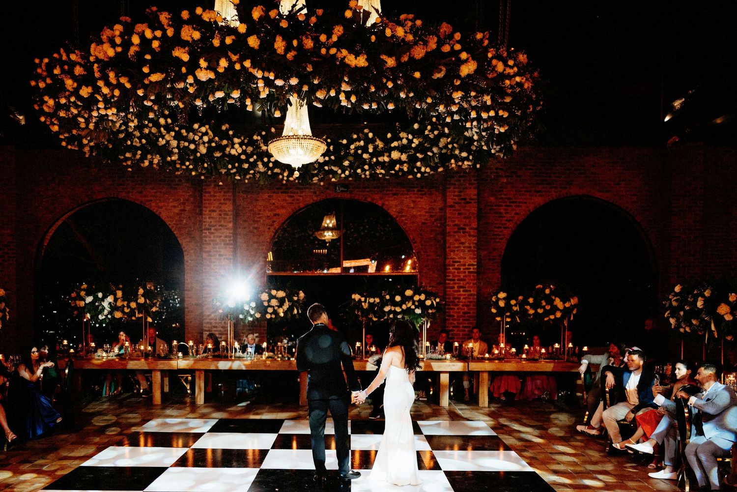 Couple shares first dance on checkered floor under floral chandelier in romantic brick venue with candlelit tables.