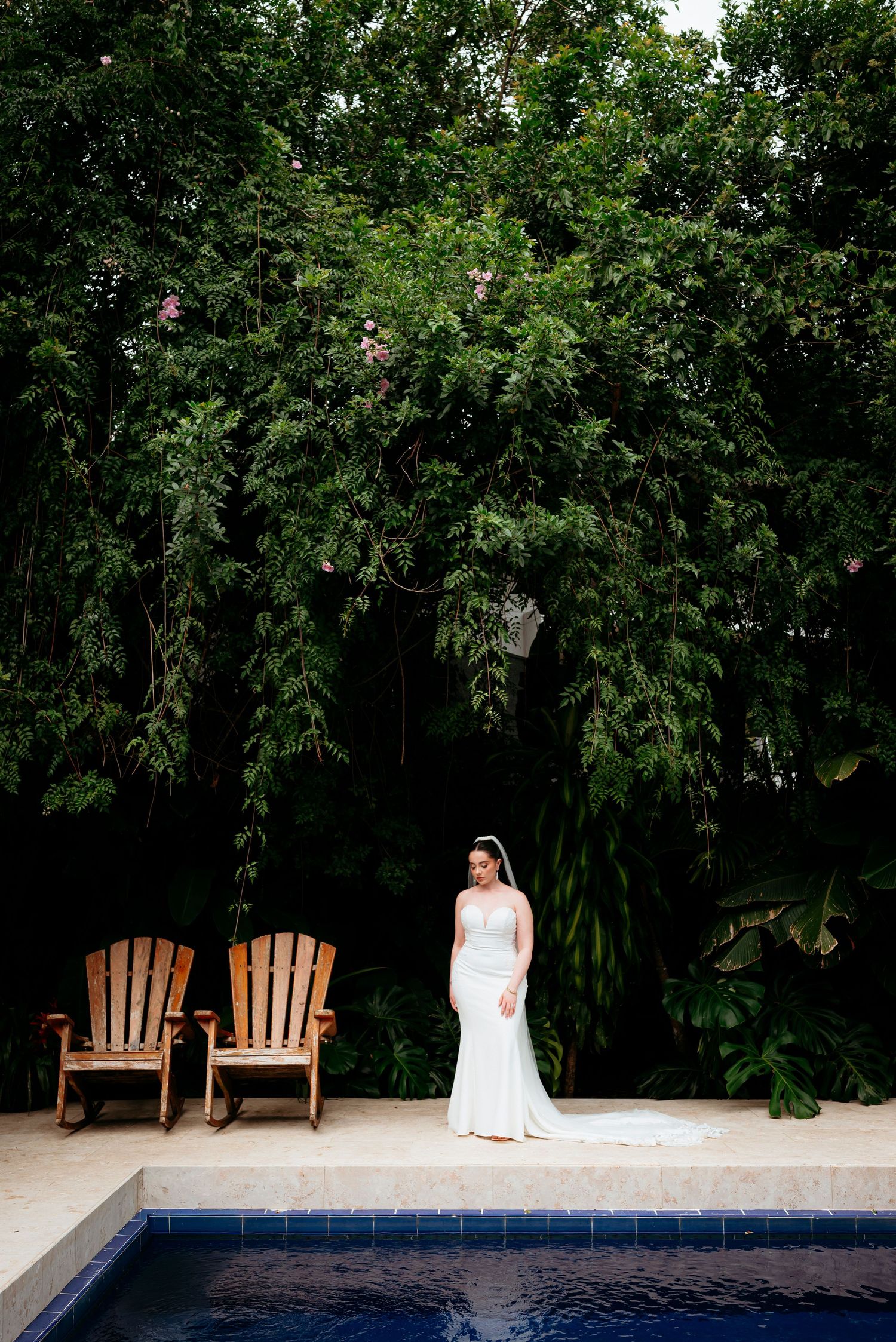 Bride in white gown stands poolside beside wooden Adirondack chairs, framed by lush green tropical trees.