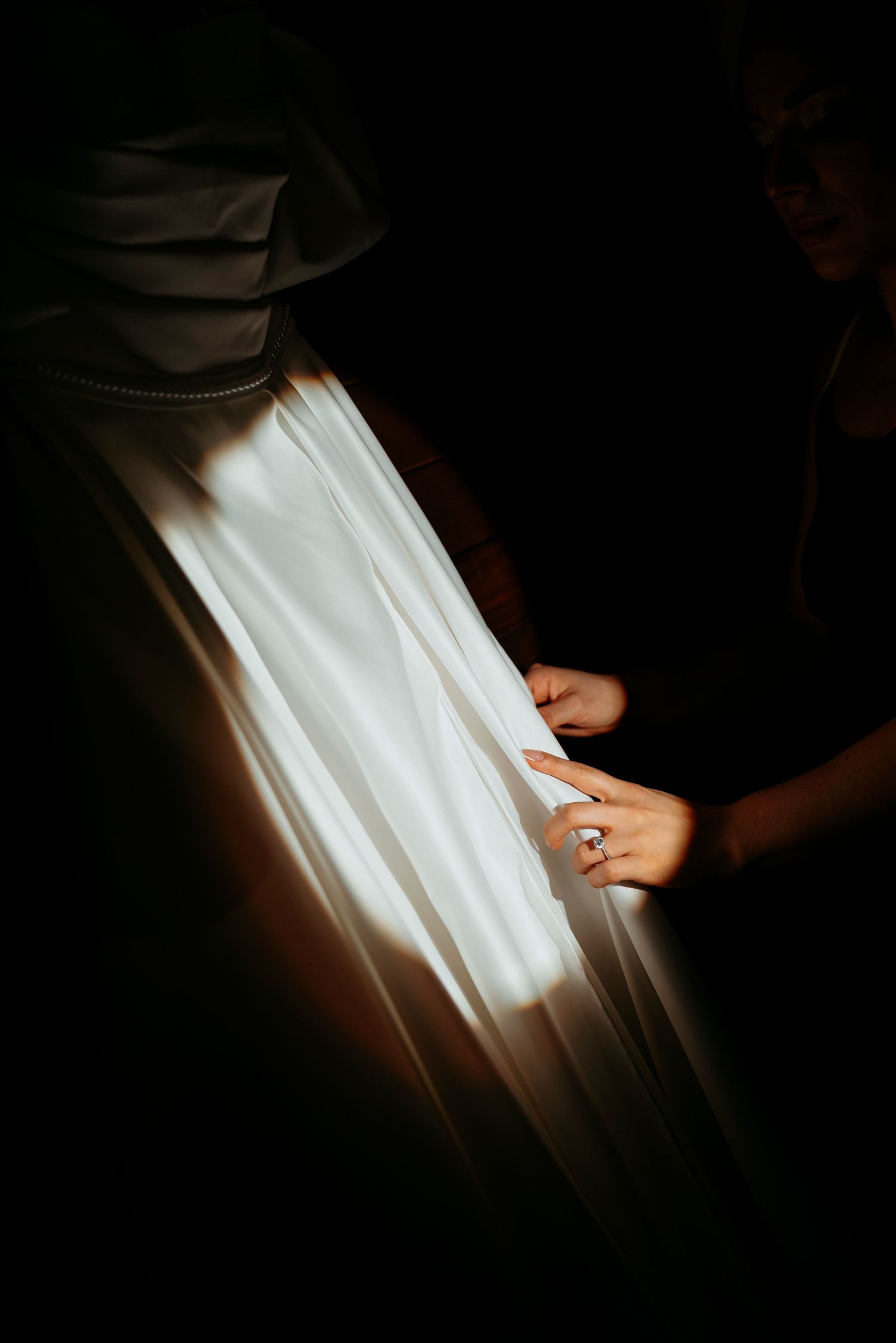 Hands with a diamond ring adjusting a glowing white wedding dress in dramatic dark lighting.