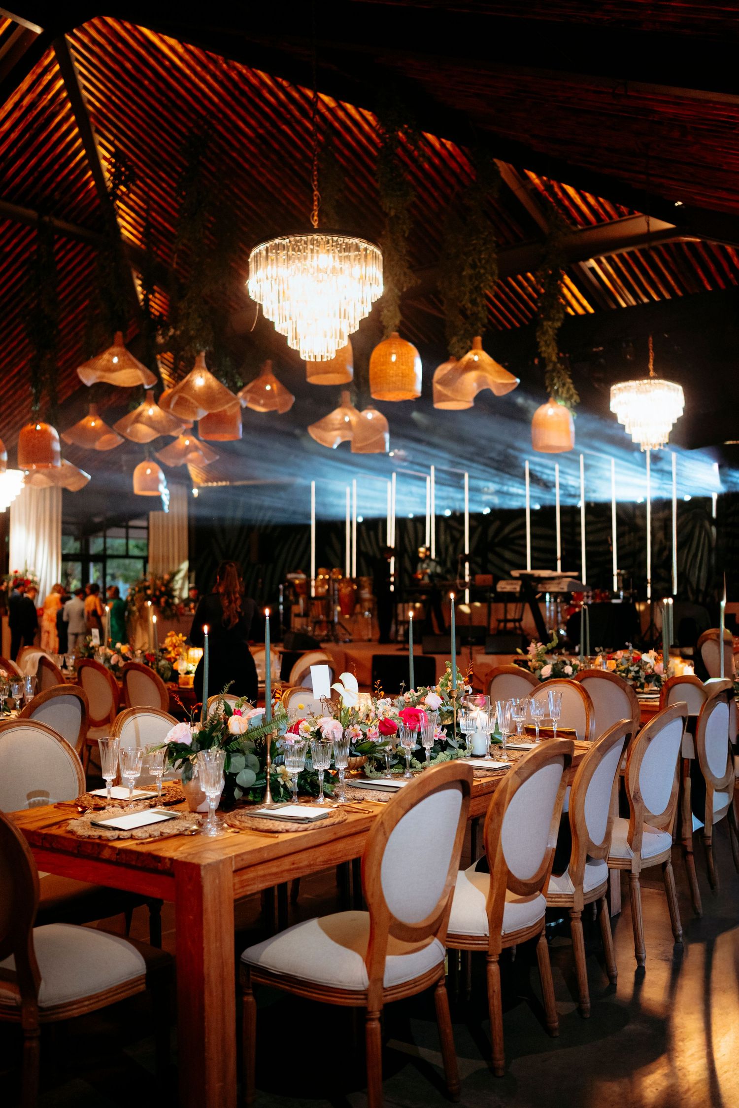 Elegant wedding reception with wooden tables, white Louis chairs, floral centerpieces, and woven pendant lights under a tropical venue.