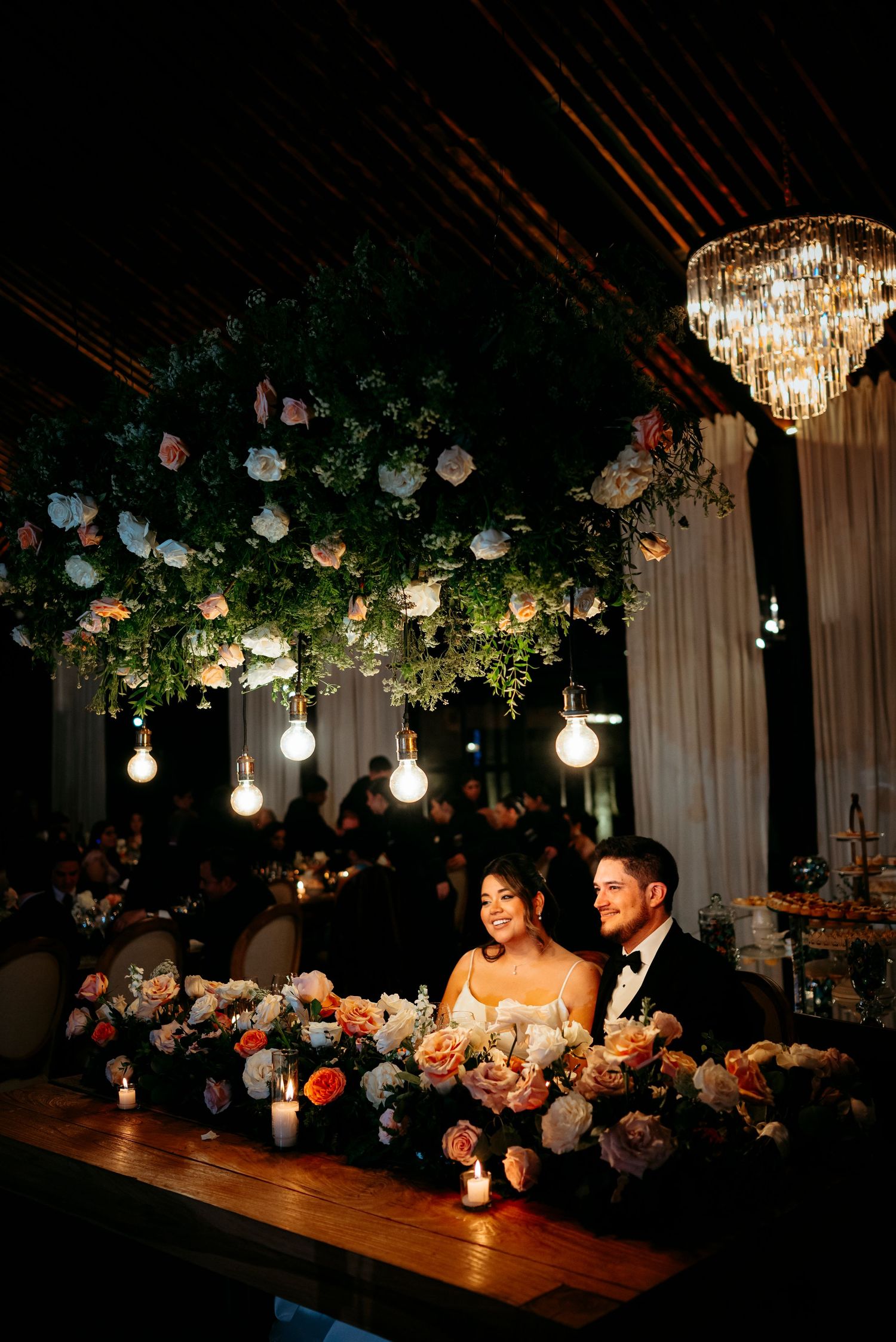 Bride and groom sit at floral-adorned sweetheart table under lush hanging flowers and glowing bulb lights at elegant wedding reception.
