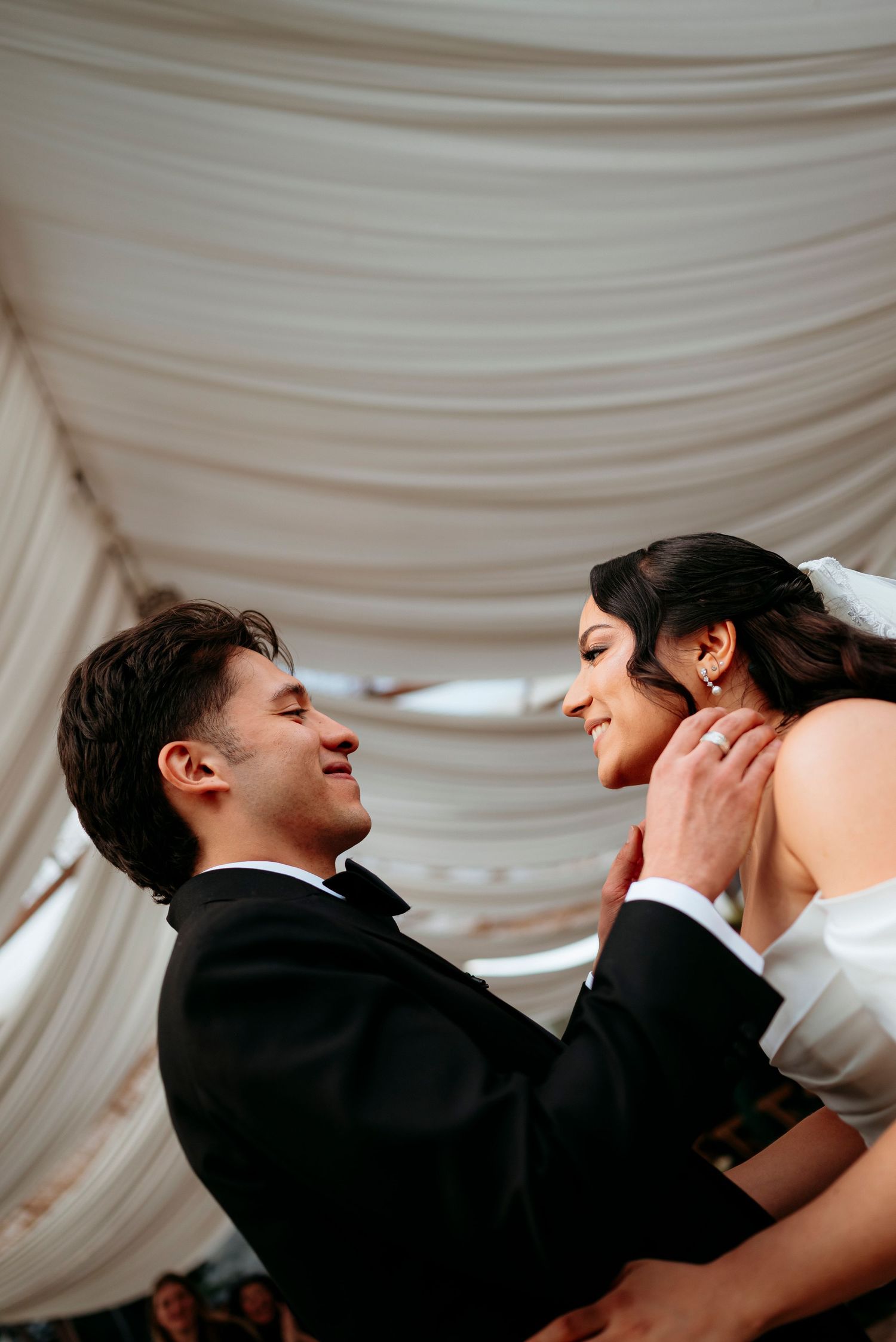 Joyful groom lifting his bride in a white gown under a draped tent at their wedding.