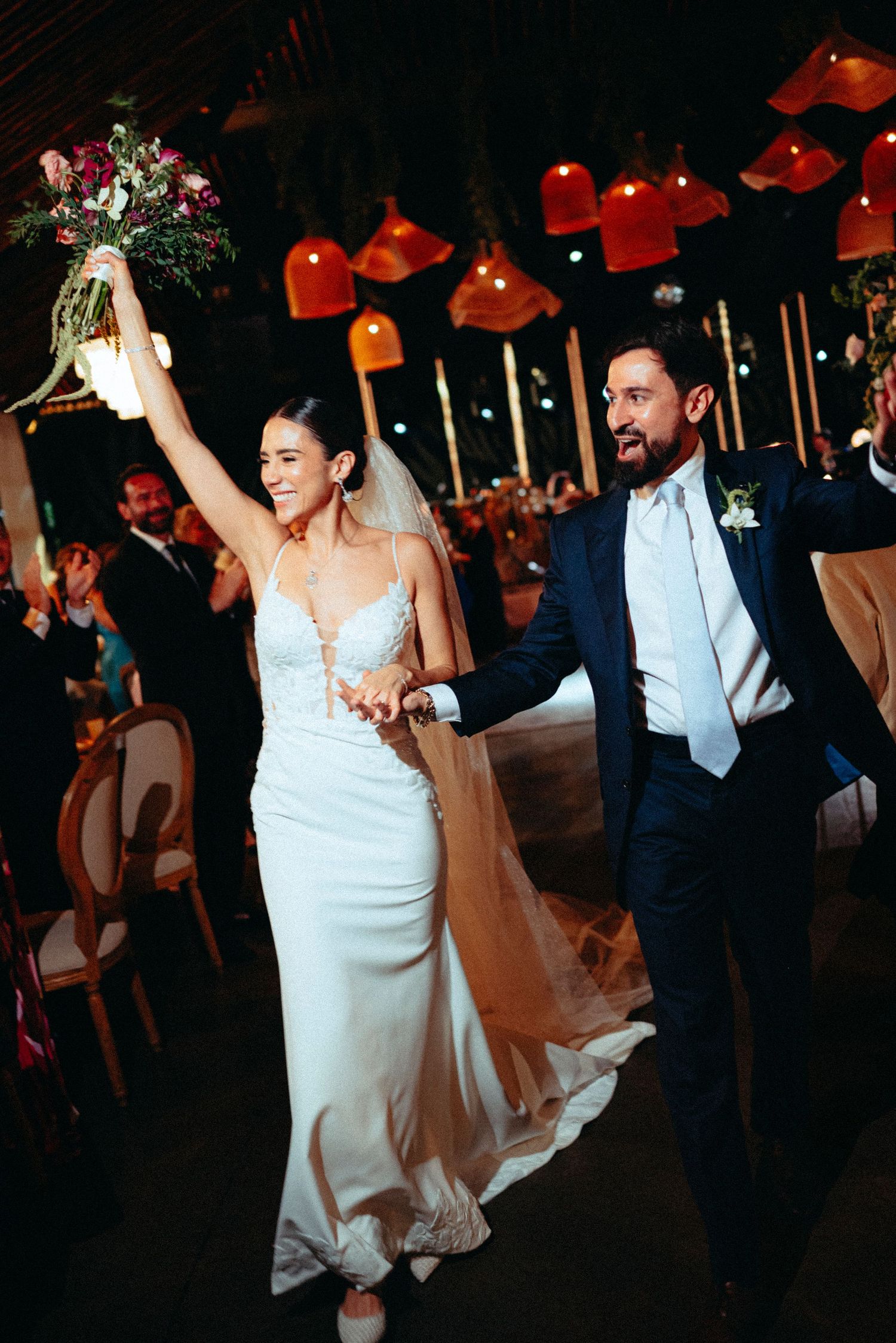 Joyful newlyweds make grand reception entrance, bride raising bouquet while groom holds her hand.