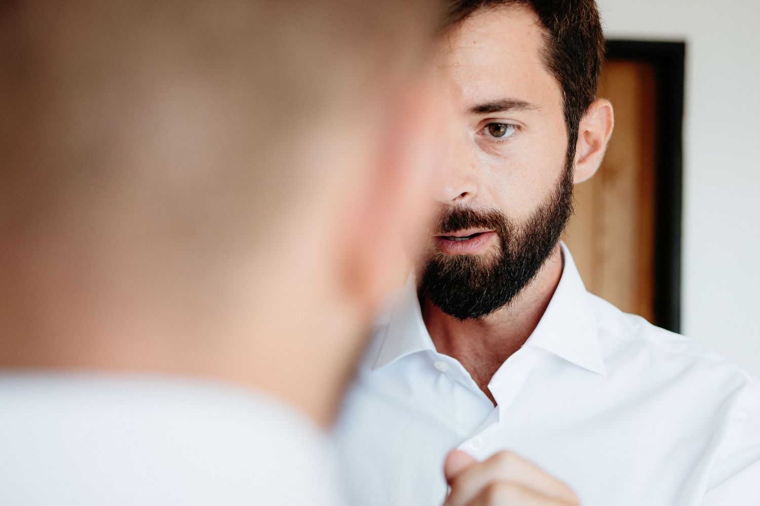 Someone in a crisp white button-up shirt adjusts their collar while looking in a mirror.