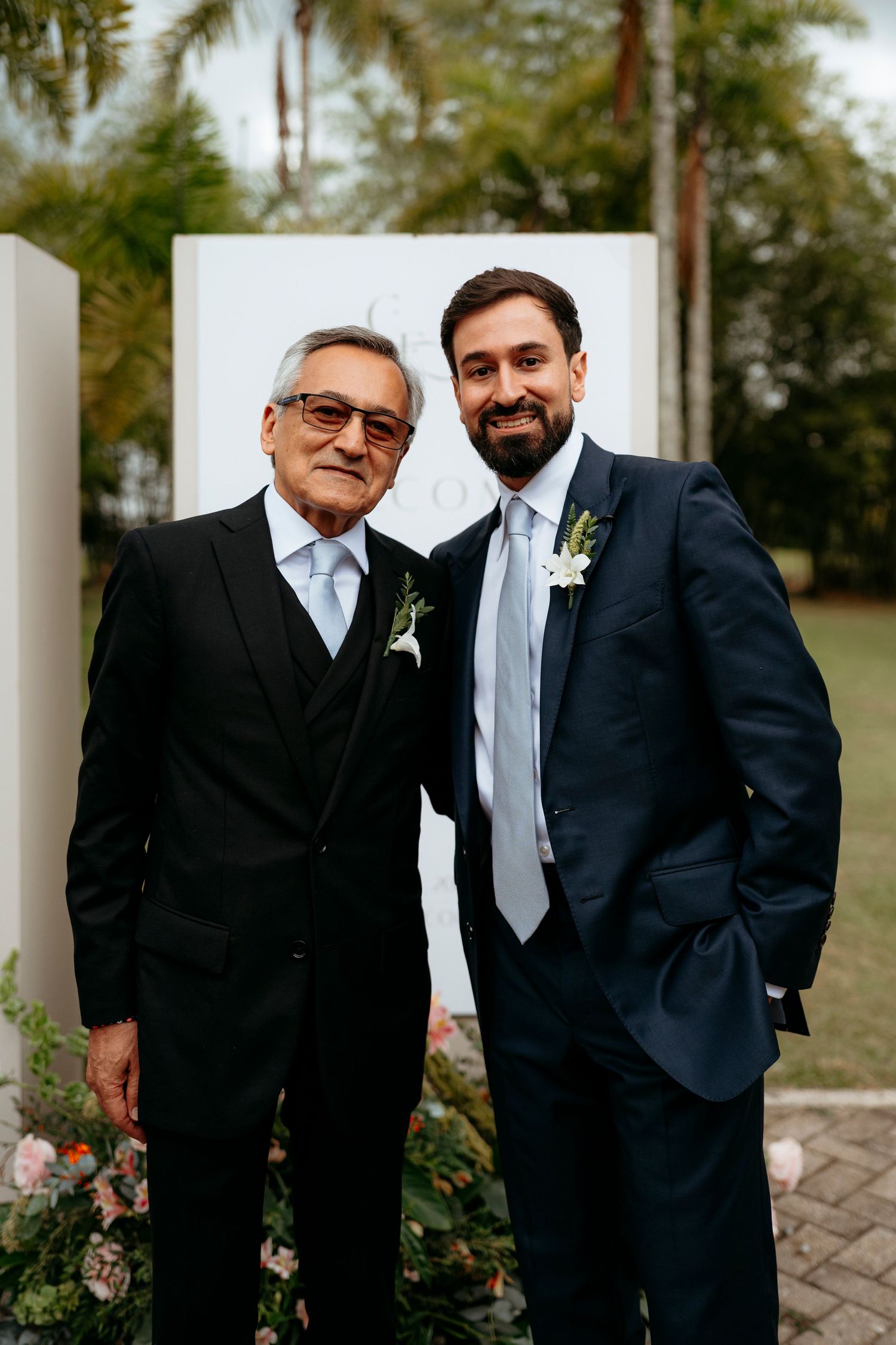 Two men in suits with boutonnieres pose outdoors at a wedding ceremony.