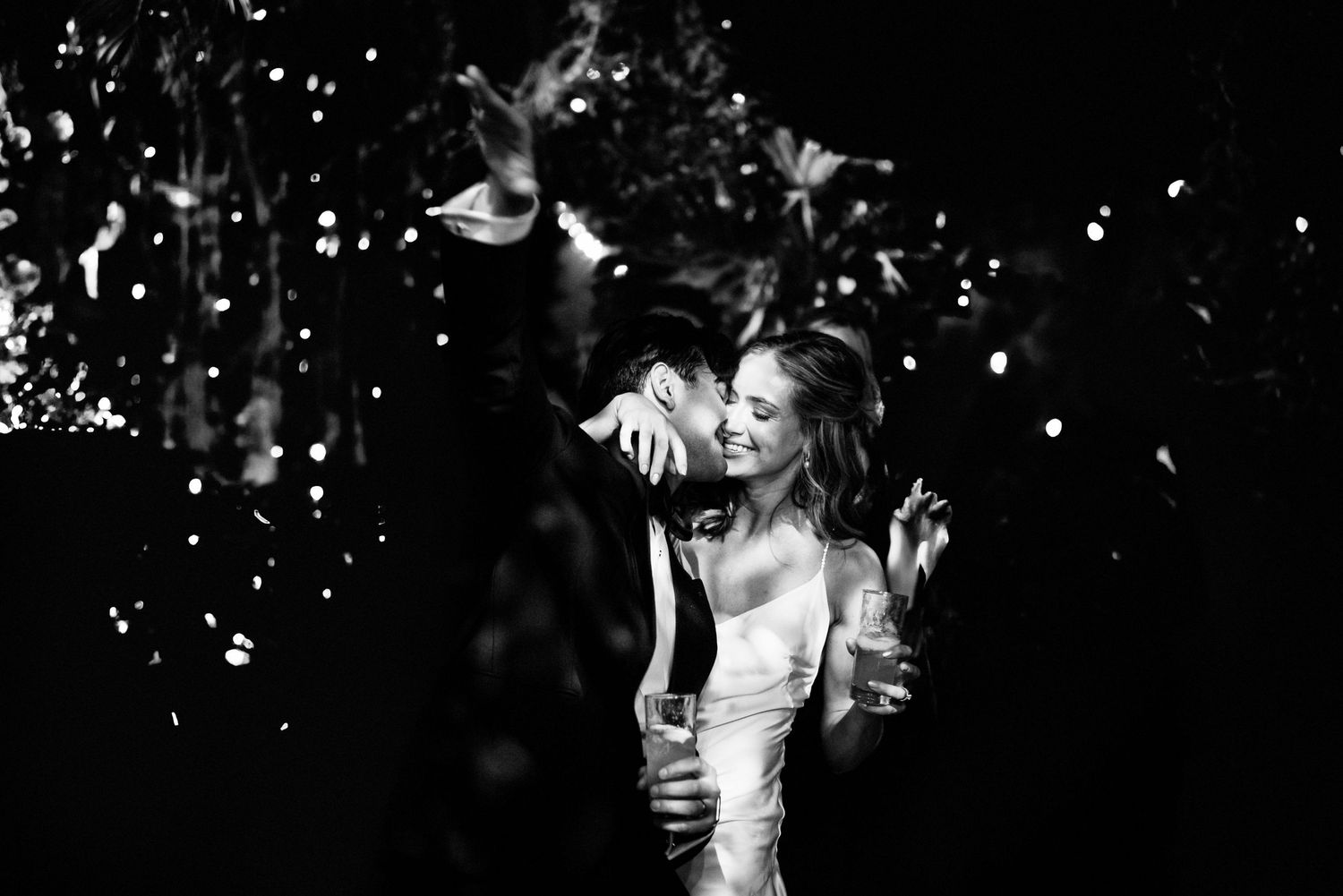 A joyful couple dances under falling confetti in dramatic black and white lighting at their wedding celebration.