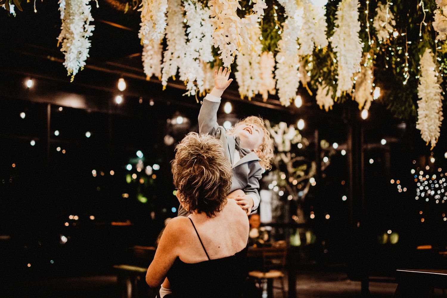 Woman in black dress holds a child reaching toward hanging white floral installation with string lights at an event.
