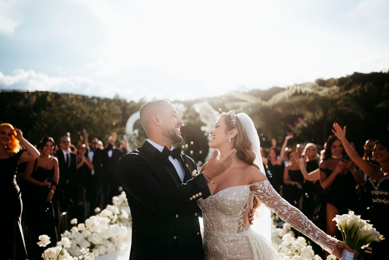 Bride and groom share a joyful moment at outdoor wedding ceremony surrounded by cheering guests and white floral arrangements.