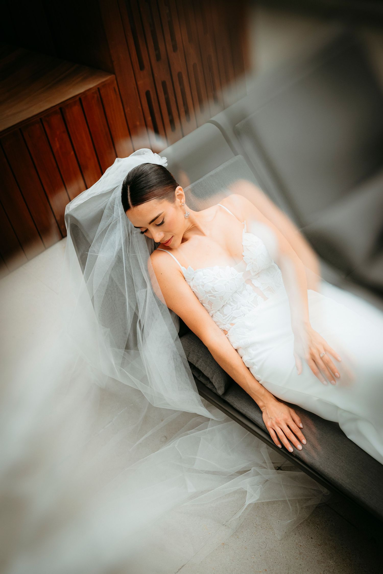 Bride in white lace gown and veil resting gracefully on a gray sofa before her wedding ceremony.