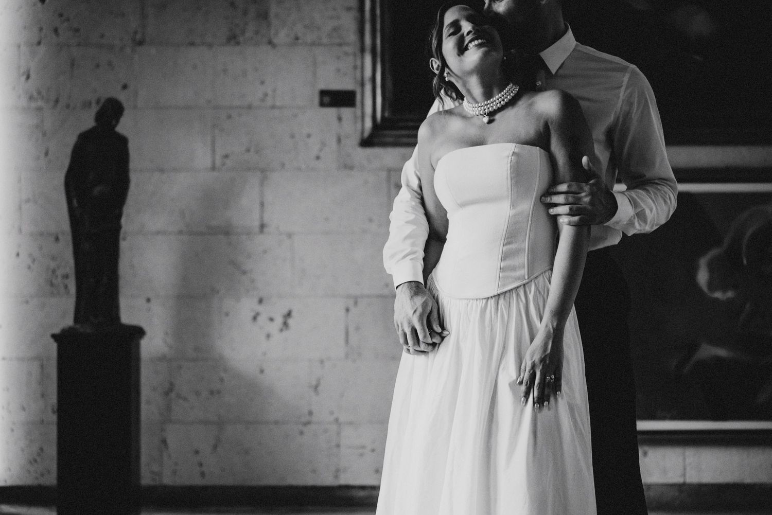 Black and white wedding photo of bride and groom embracing in elegant hall with classical statue backdrop.