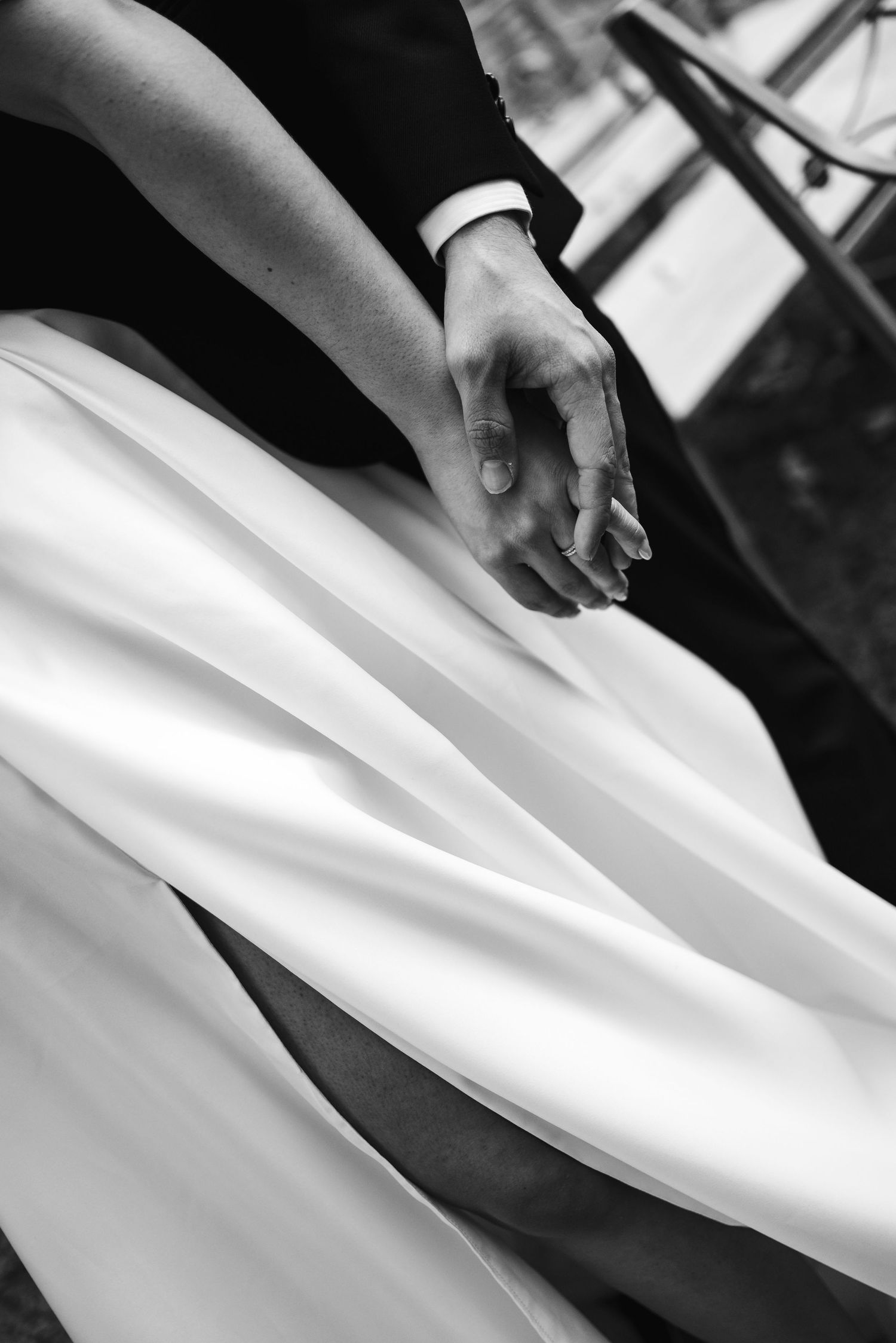 Black and white photo of a bride and groom holding hands, with flowing white wedding dress fabric in the foreground.