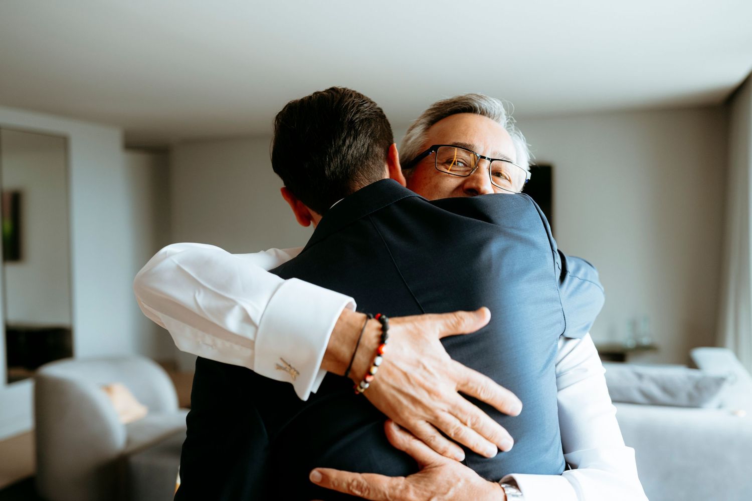 Two men in formal suits share a warm embrace, with the older man wearing glasses smiling during what appears to be a special occasion.