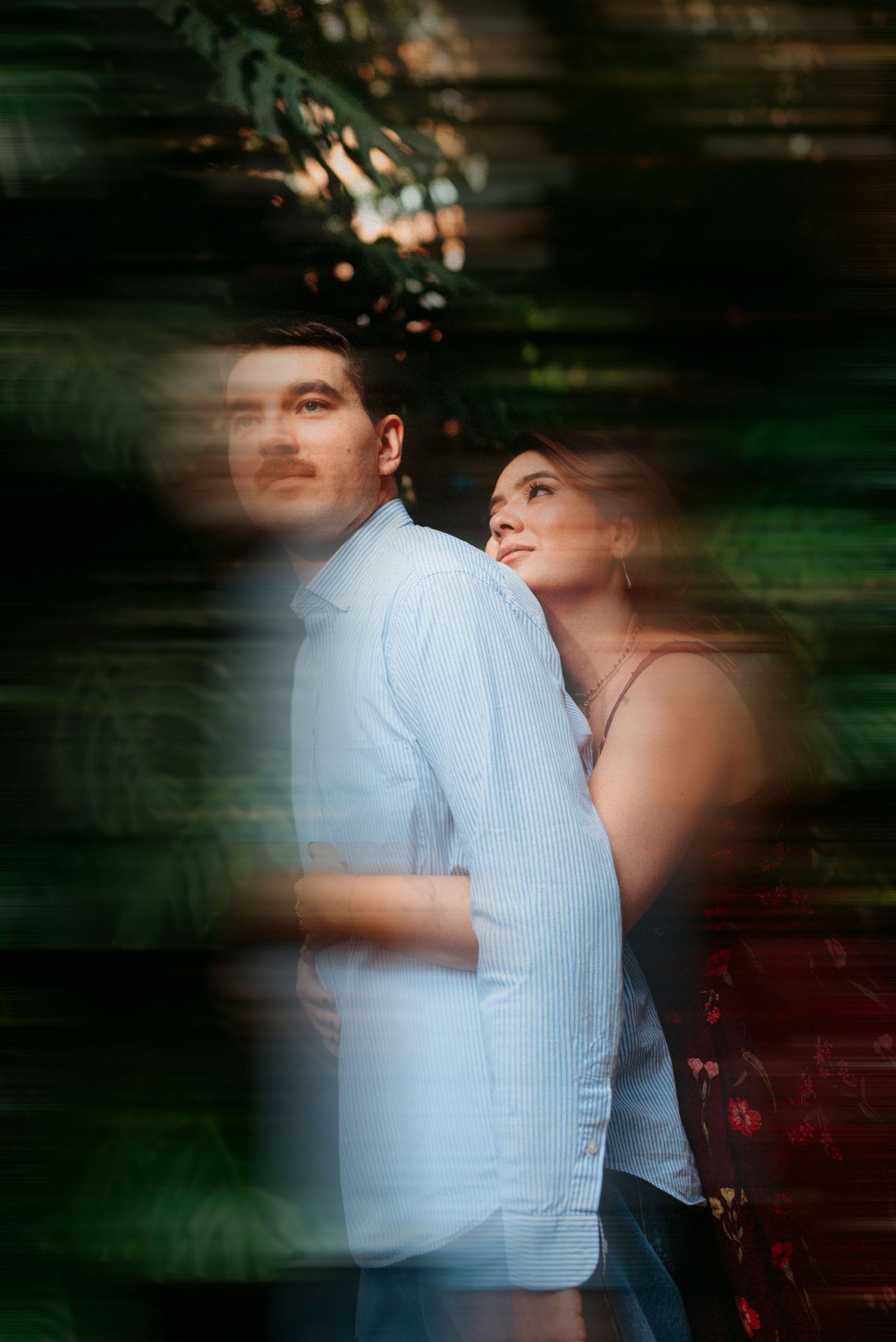 Couple embracing outdoors, man in light blue shirt, woman in dark dress, leaning together among lush green foliage.