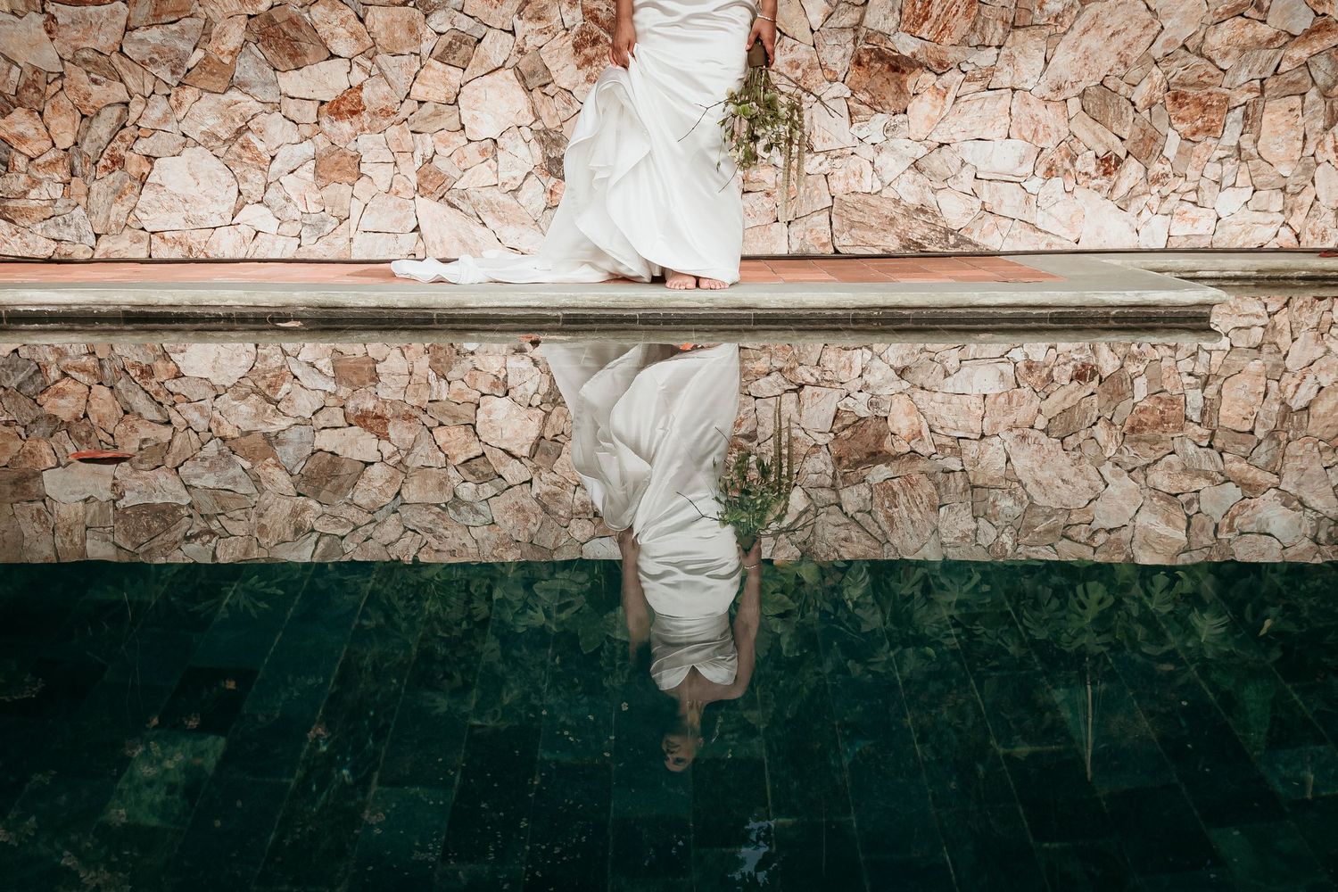 A white wedding dress hangs on a rail against a textured stone wall with a reflection in dark green flooring below.