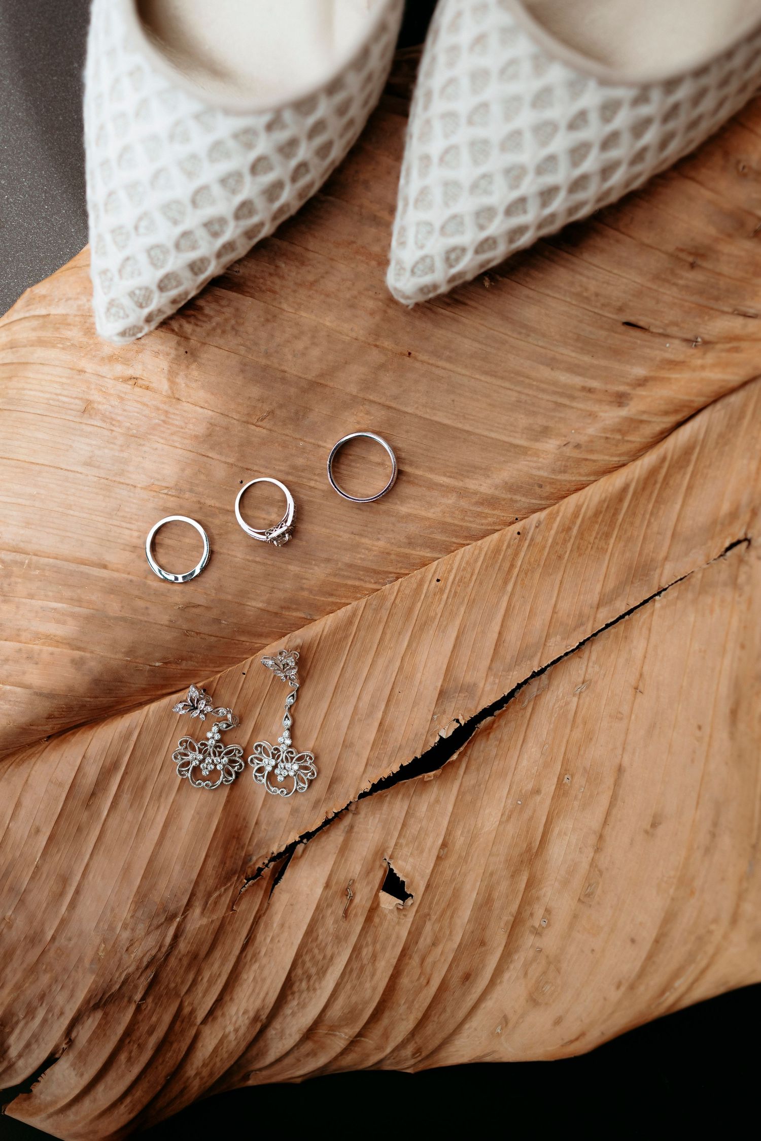 Wedding rings, diamond earrings, and white mesh heels arranged on a rustic wooden surface for a bridal flat lay.