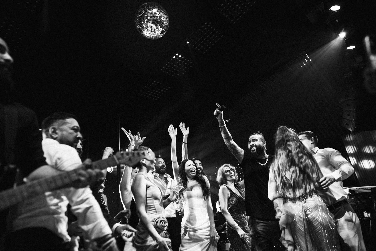 Black and white photo of energetic wedding guests dancing and cheering under a disco ball at a lively reception party.