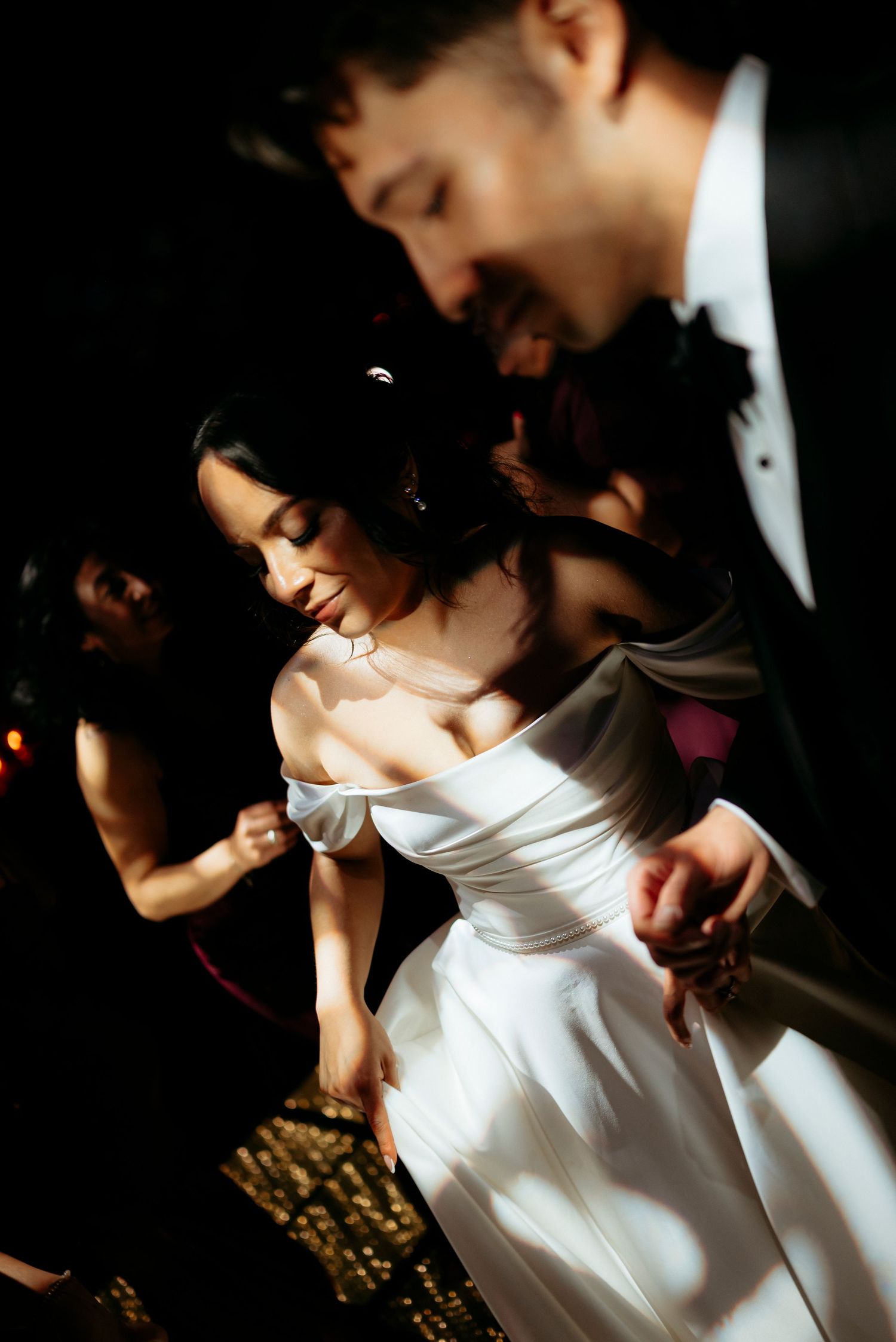 Bride in off-shoulder white gown dances with groom in tuxedo at elegant wedding reception.